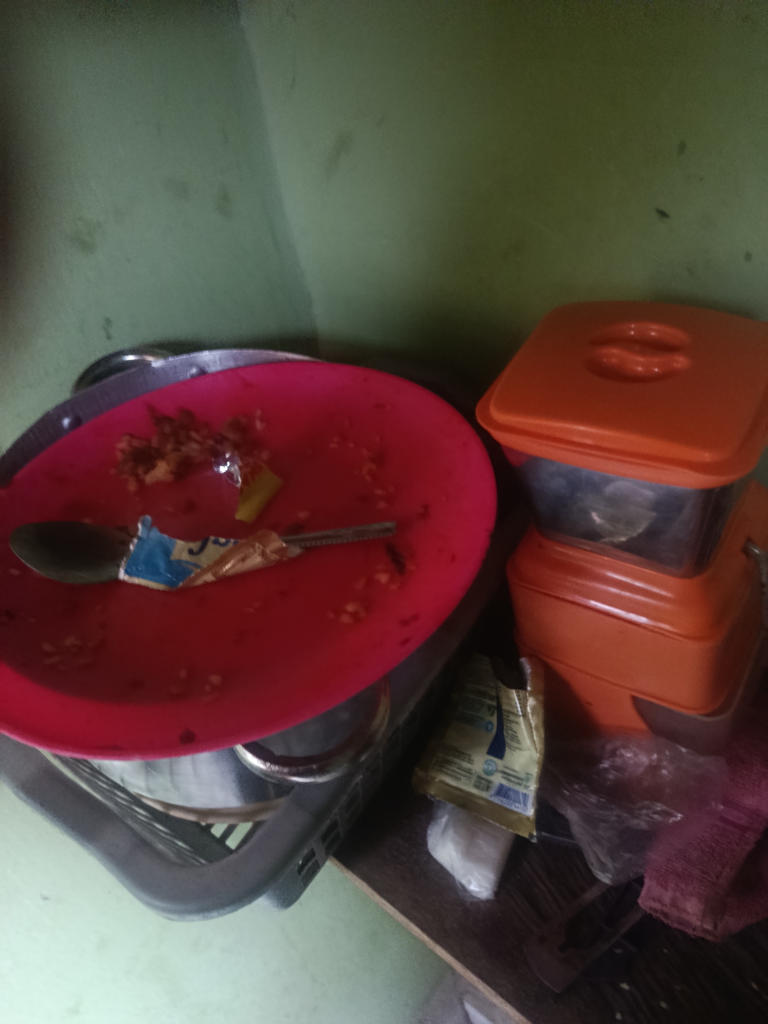 In the city of Unknown, Unknown, on a wooden shelf, there is a red plastic plate with remnants of food and a spoon. A foil wrapper with blue and yellow markings is stuck to the spoon. Beside the plate are two orange plastic food containers stacked on top of each other, with a clear container in between. To the right of the containers, there is a crumpled plastic bag and a pink mesh cloth. The background is a green wall.