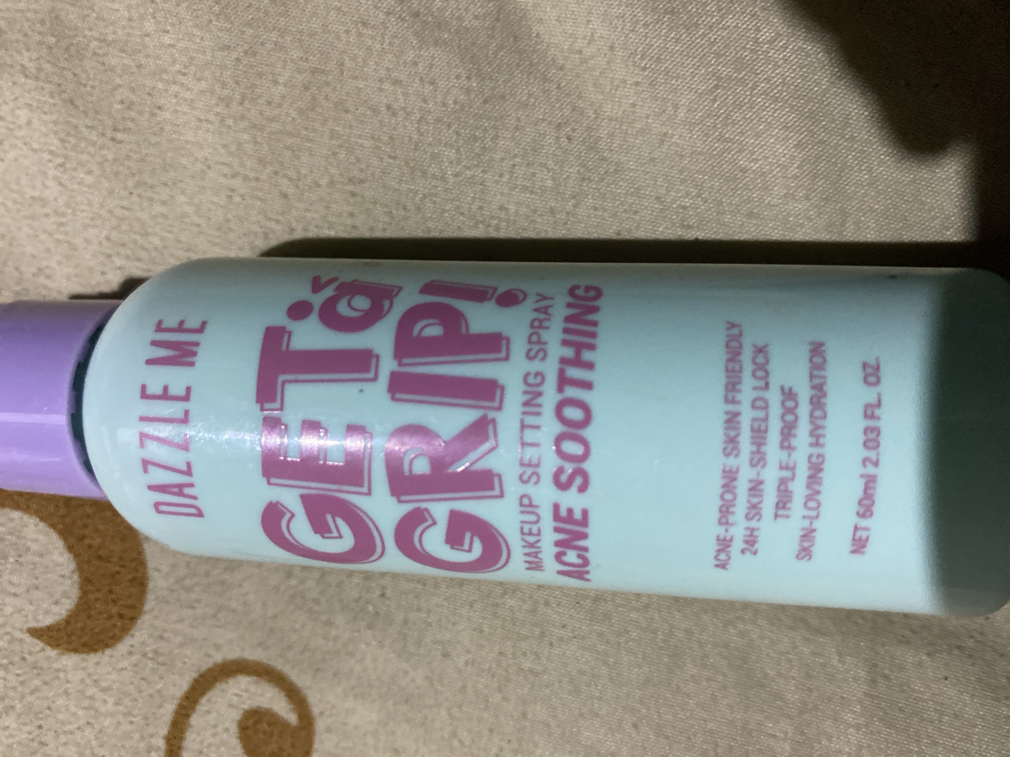 The image shows a close-up, angled view of a bottle of "DAZZLE ME GET& GRIP! MAKEUP SETTING SPRAY ACNE SOOTHING". The bottle is light blue with pink lettering. The text on the bottle reads "DAZZLE ME", "GET& GRIP!", "MAKEUP SETTING SPRAY", "ACNE SOOTHING", "ACNE-PRONE SKIN FRIENDLY 24H SKIN-SHIELD LOCK TRIPLE-PROOF SKIN-LOVING HYDRATION", and "NET 60ml 2.03 FL. OZ.". The bottle has a light purple cap. The background is a textured, light brown surface. The image does not provide any information about its location in the city of Ta', Indonesia.