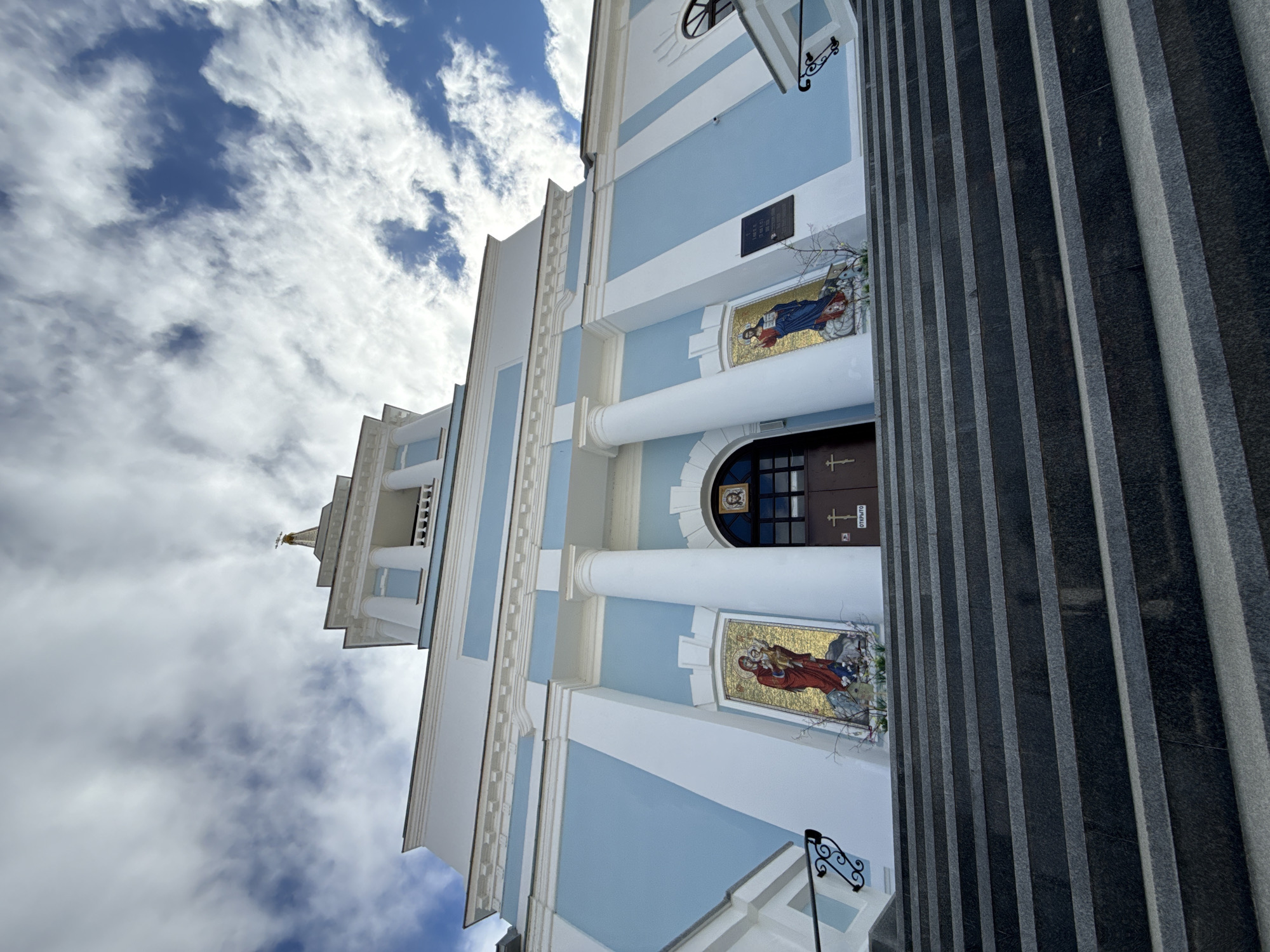 The image shows the exterior of the Orthodox Church of the Resurrection of Christ in Votkinsk, Russia. The building is painted in blue and white with neoclassical architectural elements such as columns and mosaic religious images flanking the entrance. The sky is visible in the background with scattered clouds. A tall stone staircase is also visible on the right.