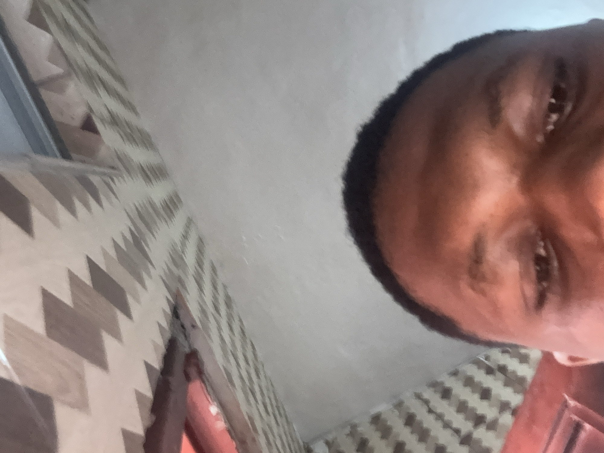The image shows a close-up, angled view of a person's forehead and eyes, with part of a ceiling visible in the background. The ceiling has a pattern of light and dark brown geometric shapes. The person's skin tone is dark, and their eyebrows are visible. The lighting appears to be artificial and casts shadows on the ceiling. The image is low resolution and slightly blurry, making it difficult to discern finer details of the person's features or the environment. There is no identifiable information within the image to suggest a specific location in Obosi, Nigeria.