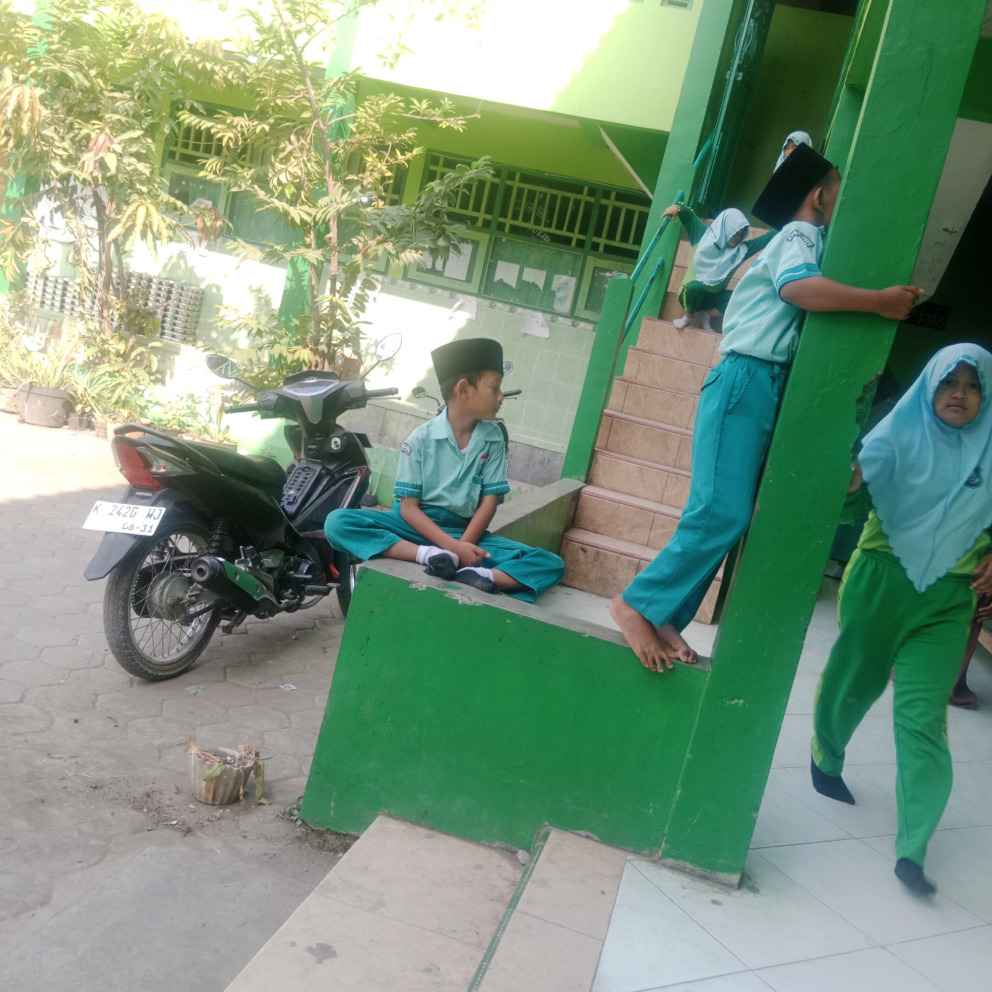 The image depicts an outdoor scene, likely on the grounds of a school, in Tegowanu Kulon, Indonesia. A young boy wearing a light blue uniform and a black peci (cap) is sitting cross-legged on a low, green wall, facing to his right, with his back to a black motorcycle parked behind him. The motorcycle has a license plate reading "K 2420 WJ" and "06-31".
To the right of the boy, a green staircase ascends. Several other children are visible on or near the stairs, also in similar light blue and green uniforms. One child, wearing a light blue hijab, is partially visible at the top of the stairs, and another child in a light blue hijab and green uniform is walking down the stairs. Two other children are further up the stairs, one leaning on the railing.
The environment features lush green foliage, suggesting a tropical or subtropical climate. The building in the background is painted green, with windows that have green bars. The ground is paved with interlocking concrete tiles. A small, weathered pot containing dry leaves sits on the tiled ground near the motorcycle. The overall scene appears to be a casual moment, possibly during a break or between activities at a school.