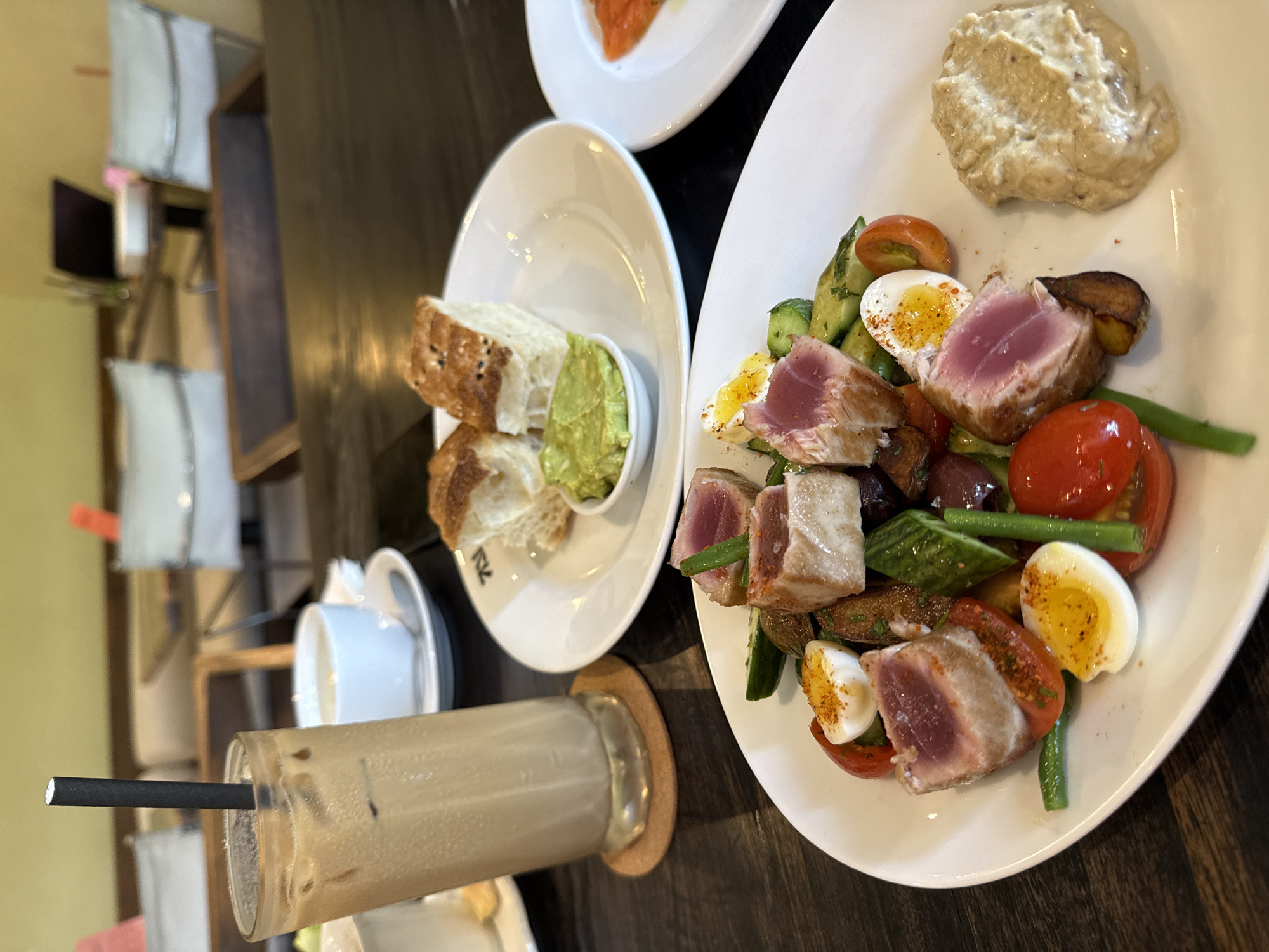 This image captures a close-up, high-angle view of a diverse meal spread on a dark wooden table in what appears to be a cafe or restaurant in North Kuta, Indonesia. The scene suggests a relaxed dining experience for at least two people.
In the foreground and center, a large white, elongated plate holds a vibrant seared tuna salad. The tuna is cooked rare, with a distinct pink center, and is seasoned with spices. The salad features halves of hard-boiled eggs sprinkled with paprika, cherry tomatoes, green beans, cucumber slices, black olives, and roasted potato pieces. A dollop of a creamy, light-colored dip, possibly hummus or a similar spread, is placed at the top of the plate.
To the left, another white plate contains slices of crusty bread with black sesame seeds, served alongside a small white bowl of mashed avocado or guacamole. Partially visible behind this plate is a portion of a third white plate holding what appears to be sliced salmon.
An iced coffee or latte in a tall glass, complete with a black straw, is positioned in the lower-left corner, resting on a round cork coaster. A white ceramic teacup and saucer are also visible in the background, slightly blurred. The background of the image is softly out of focus, showing glimpses of chairs with white cushions and shelves or partitions, indicating an indoor dining establishment.
The overall lighting suggests it is daytime. On the plate with the bread and avocado dip, stylized letters resembling "ZK" or "NK" are visible near the rim.