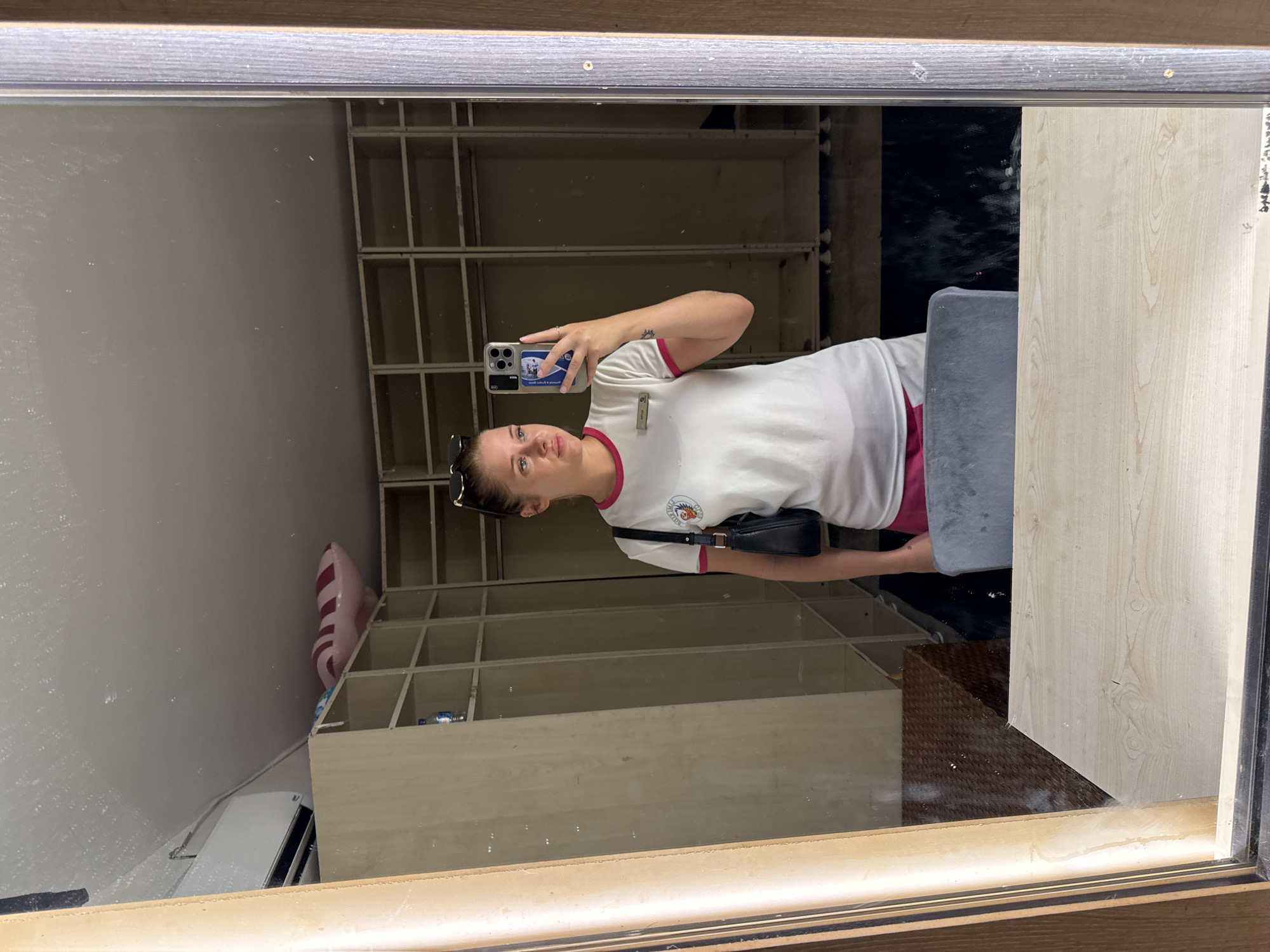 The image shows a young woman taking a mirror selfie in what appears to be a dressing room or storage area. She is wearing a white t-shirt with pink trim and pink shorts. She has sunglasses on top of her head and a black purse slung over her shoulder. In the reflection, we see a row of empty shelves and an air conditioning unit mounted on the wall. The room is sparsely furnished, with a few items reflected in the mirror, including what looks like a brown woven basket and a grey chair. The overall impression is one of a functional, perhaps temporary, space. The location is identified as Aksu, Turkey.