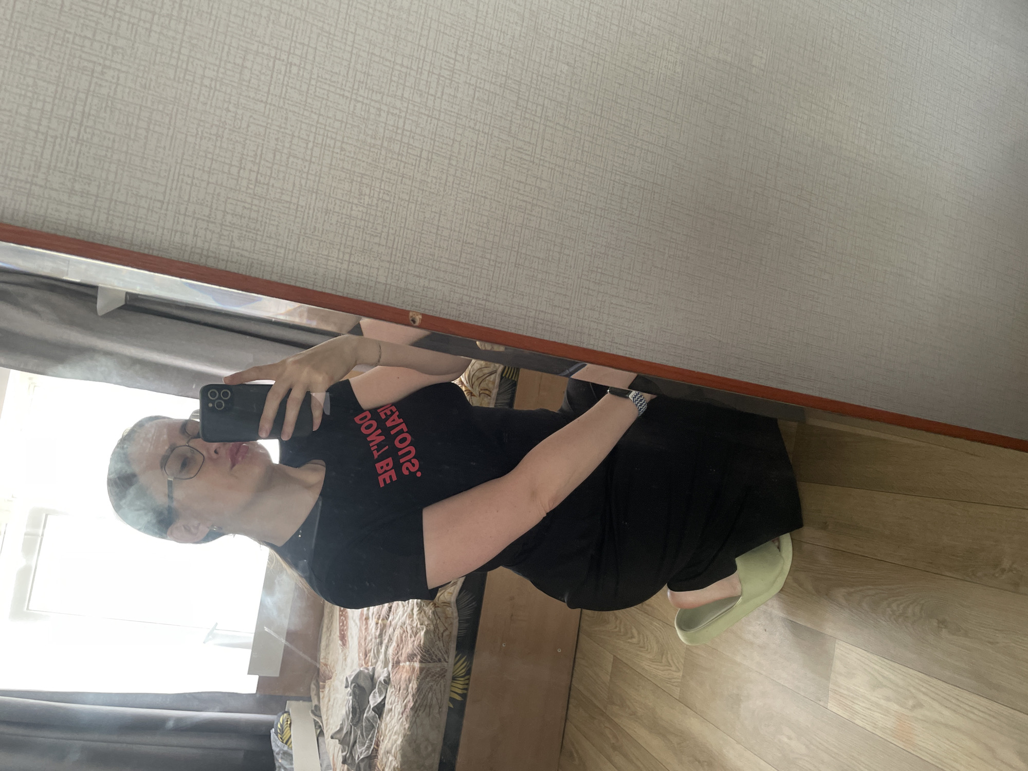 A young woman with glasses is taking a mirror selfie in a room. She is wearing a black t-shirt with pink text that reads "DON'T BE JEALOUS" backwards, and a long black skirt. She is kneeling on a wooden floor, and her feet are clad in light green slippers. In the background, a bed with patterned bedding is visible. The room appears to be a bedroom, with a window providing natural light. The mirror reflects the woman, her phone, and the room behind her. The overall scene suggests a casual, private setting within a home in Syrokovo, Russia.