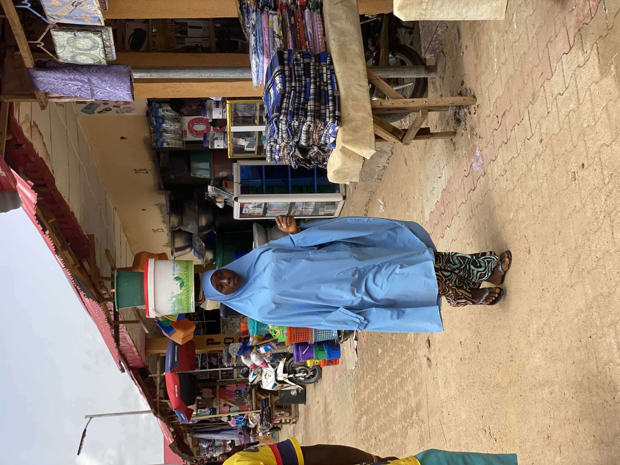 A woman wearing a blue robe and head covering walks along a street in Kafanchan B, Nigeria. She is carrying several buckets stacked on her head. The street is lined with shops displaying merchandise, and a motorcycle is parked nearby. The ground is paved with reddish-brown and light gray rectangular bricks. The sky is overcast.