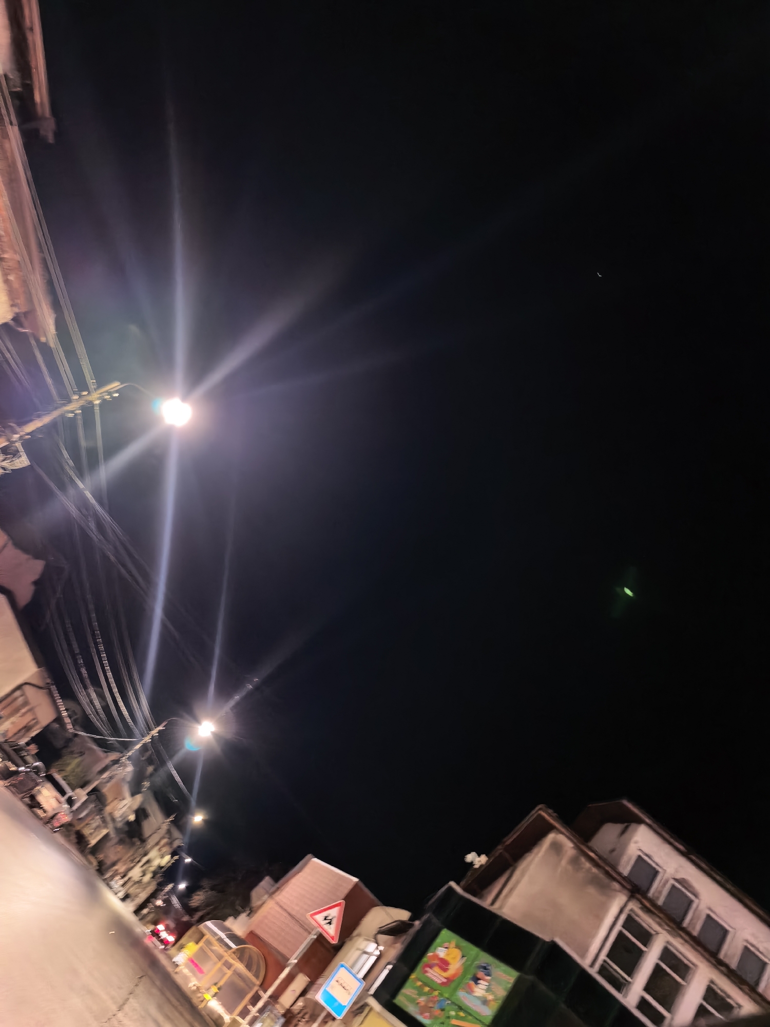 The image depicts a nighttime street scene in an urban environment. Streetlights illuminate the scene, casting bright rays across the sky. Buildings are visible on either side of the street, with their architectural details partially obscured by the darkness. Power lines stretch across the upper portion of the image. Traffic signs are present on the right side of the street. A crescent moon is faintly visible in the dark sky. There are no people visible.