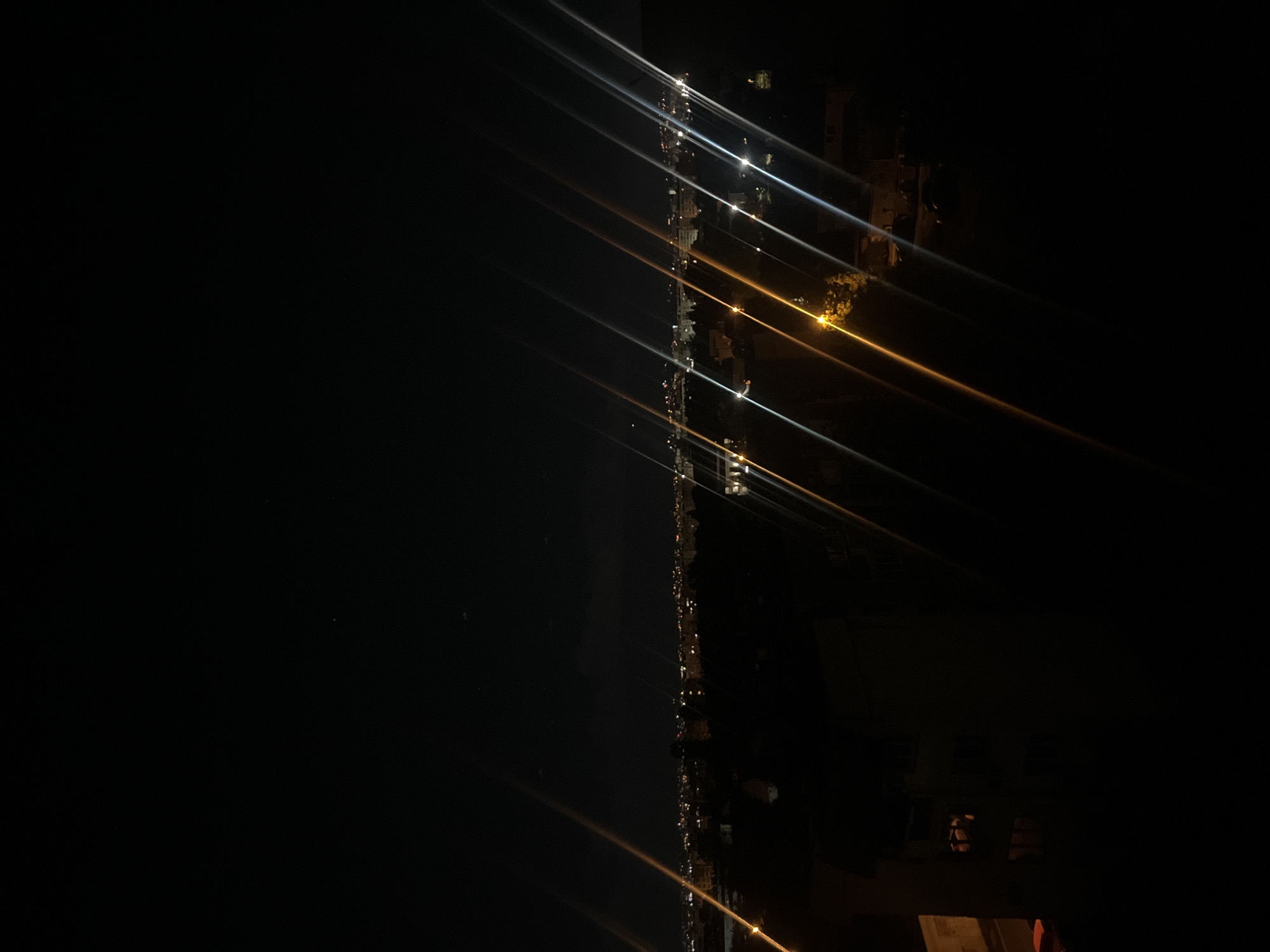 At night, a long line of city lights appears to run vertically through the center of the frame, with an impression of being reflected in a dark body of water. The lights vary in color, with some appearing white and others yellow. Streaks of light extend outwards from the individual points of light, adding a sense of movement and distortion. The rest of the frame is dominated by darkness, suggesting that the scene is a nighttime urban view. Without additional information or visual cues, it is impossible to confidently pinpoint the exact location in Sofia, Bulgaria, but based on the urban lights, it could be near a lake or reflection of a major street with buildings around it.