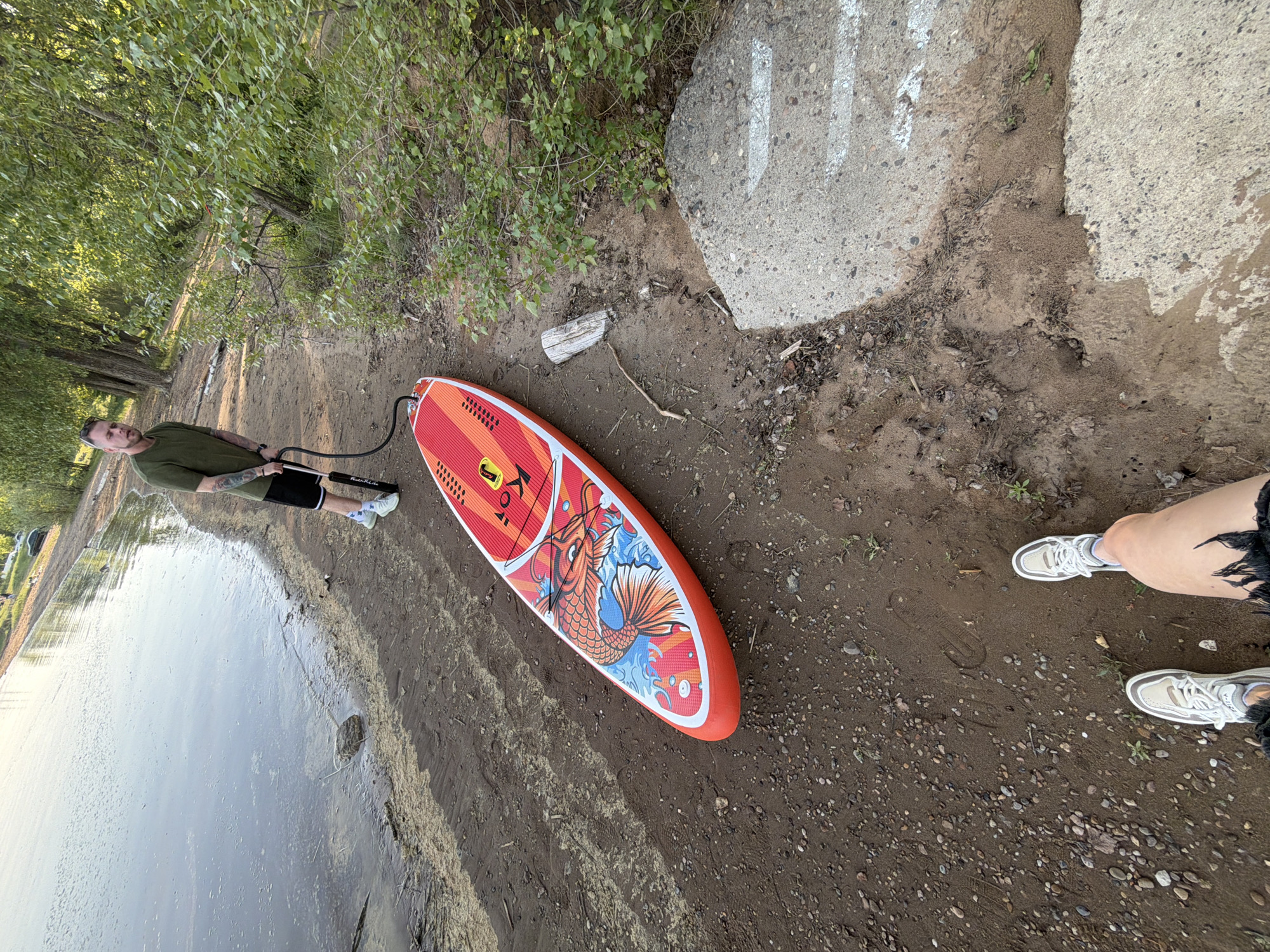 A man is inflating a paddleboard on a shoreline. The paddleboard is orange and has a koi fish design. The shoreline is muddy with pebbles and small rocks. The water is visible to the left. There is a person's legs and shoes on the shoreline. Bushes and trees line the shore. The GPS coordinates of the image are unavailable, and therefore I cannot provide the location in the city of Noviy, Russia.