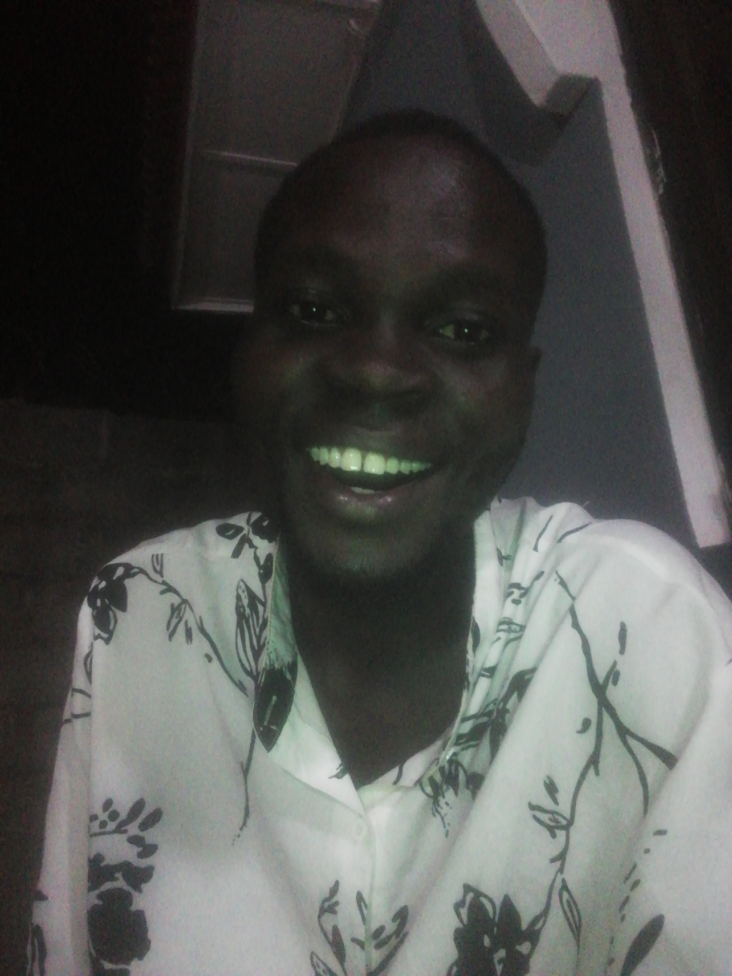 A person with dark skin is smiling at the camera. They are wearing a light-colored button-down shirt with a dark floral pattern. The background is dark, but a corner of a white-painted wall is visible in the upper right. The location is suggested as Ikorodu, Nigeria.