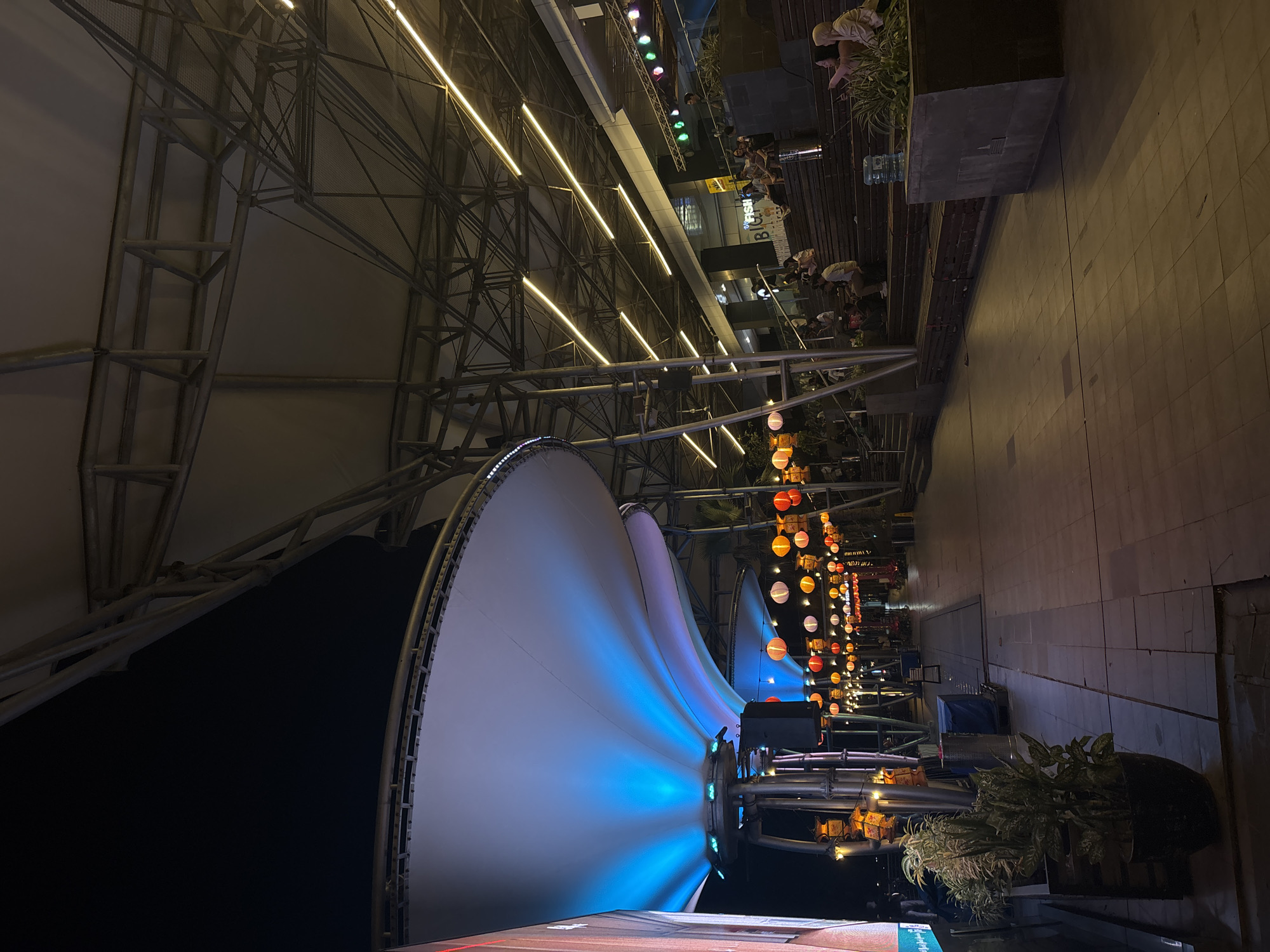 The image depicts an outdoor setting in North Jakarta, Indonesia, at night. A large, white, sail-like structure, illuminated with blue light, dominates the left side of the frame. The structure is supported by a steel framework. Strings of orange lanterns stretch into the distance along the right side of the frame. People are seated at tables on a wooden deck behind the lanterns. The ground is paved with rectangular tiles. Planter boxes with greenery are visible along the edge of the paved area.