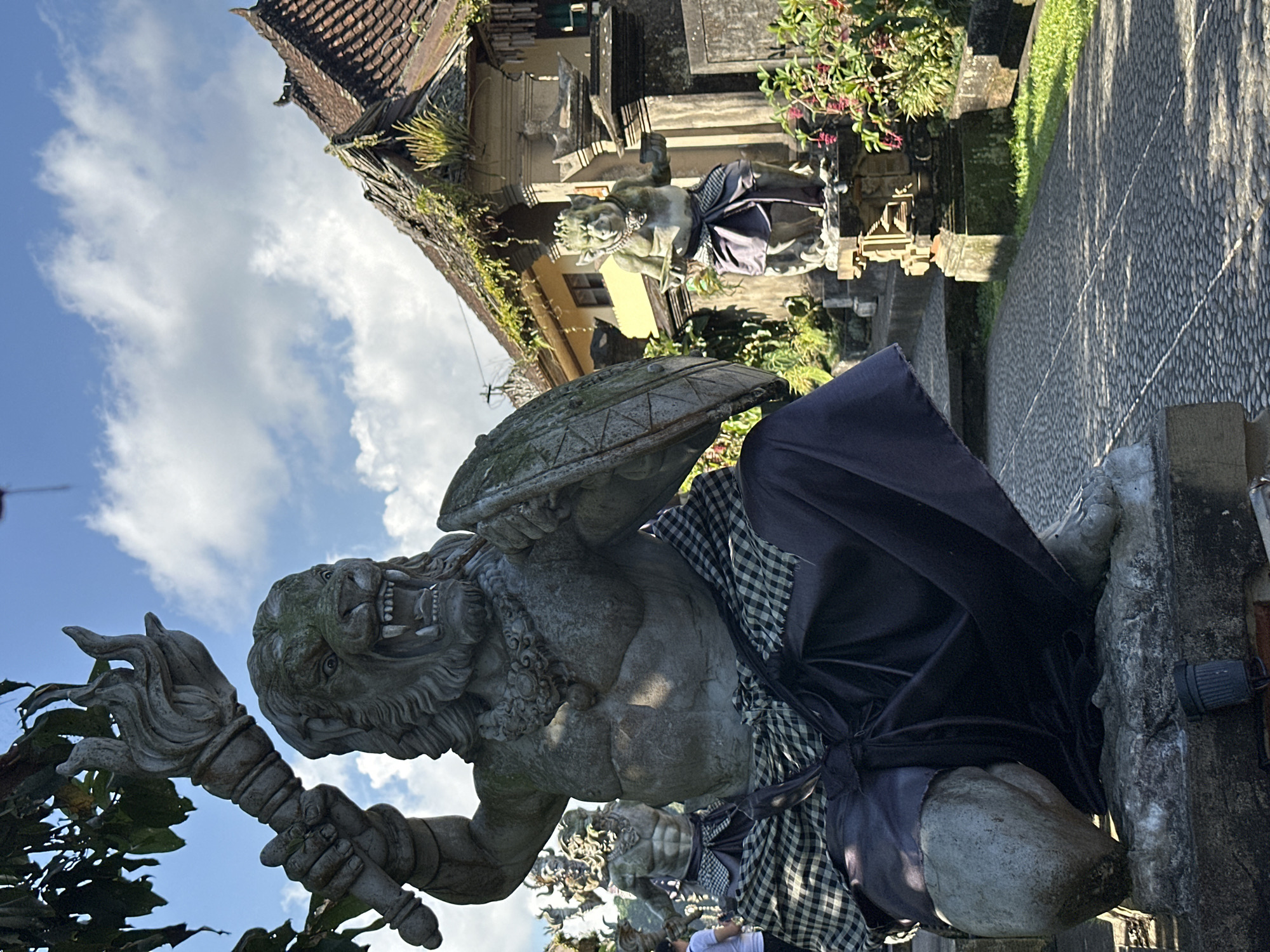 This is a vertical, eye-level, outdoor photograph captured in Bangli, Indonesia, showcasing a pair of Balinese temple guardian statues. In the foreground, a large, imposing stone statue of a fearsome creature with a lion-like head and a muscular human torso kneels on a stone pedestal. The statue holds a flaming torch in its raised right hand and a shield in its left. It is adorned with a patterned fabric sarong around its waist. Behind this statue, further into the scene, another, smaller stone statue of a grotesque, pig-like creature with a crown and a sarong also kneels. The statues are situated along a paved pathway constructed of small, grey cobblestones. To the right, the corner of a building with a traditional tiled roof, partially overgrown with greenery, is visible. The environment is sunny with a bright blue sky dotted with white clouds. Lush green foliage and flowering plants surround the statues and the pathway. The overall scene depicts a cultural and spiritual setting, characteristic of Balinese temples or public spaces with religious art.