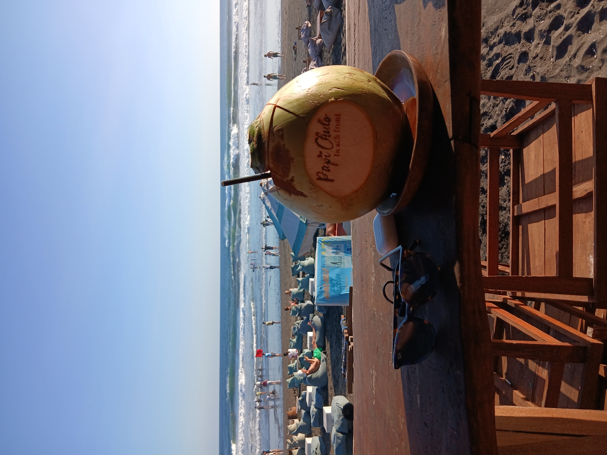 The image depicts a close-up view of a table at a beachside establishment, with a whole coconut drink placed on a wooden plate in the foreground. The coconut has "Papi Chulo beach front" carved into its surface and a straw inserted. Next to it, a pair of sunglasses and a white earbud case rest on the table. The table is made of dark wood, with a wooden structure beneath it visible. In the background, the sandy beach of North Kuta, Indonesia, is populated with people relaxing on beanbag chairs and walking along the shore. The ocean, with moderate waves, stretches to the horizon under a clear blue sky. The scene suggests a leisurely atmosphere at a beachfront cafe or bar.