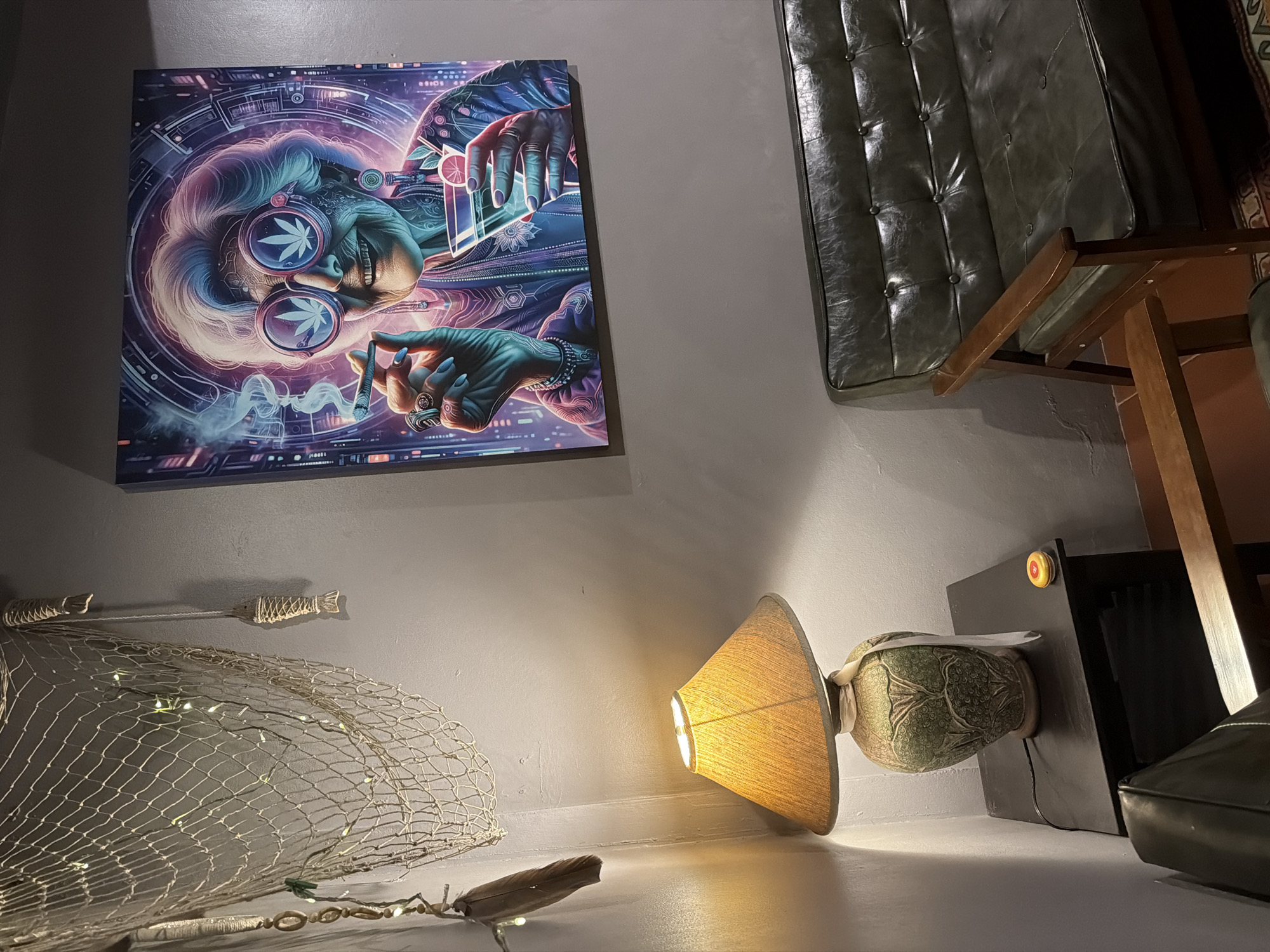 This media file depicts an indoor scene, likely a room, with an artistic painting as a prominent subject. The painting, hanging on a grey wall, is a stylized portrait of an elderly person with distinctive glasses, featuring cannabis leaf symbols. The artwork is rendered in vibrant blues, purples, and pinks, with a futuristic or psychedelic aesthetic, showing hands holding what appears to be a cigarette and cards.
In the foreground, a fishing net is draped, adorned with small, white fairy lights. A single feather rests on a surface below the net, also illuminated by faint lights. To the right, a lamp with an orange shade casts a warm glow. Beside the lamp, a decorative, green patterned vase-like object is visible, resting on a dark surface. Behind this, part of a dark leather armchair or sofa is visible, along with the legs of a wooden chair or table.
The lighting suggests it might be nighttime or a dimly lit interior. The overall mood evoked by the combination of the art, the decorative elements, and the lighting is eclectic and perhaps bohemian. There is no visible text within the image.