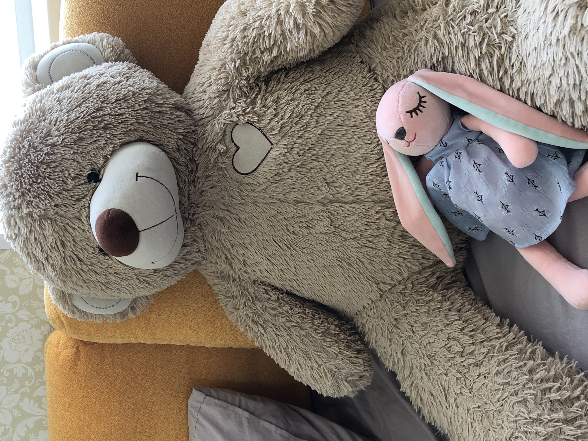 A close-up shot displays a large, stuffed, light brown teddy bear with a white heart patch on its chest, resting on a mustard-colored cushion and grey bed linen. A plush pink rabbit toy wearing a blue dress with a white pattern is placed on the teddy bear. The setting appears to be indoors. The city of Noviy, Russia is not discernible from the image.
