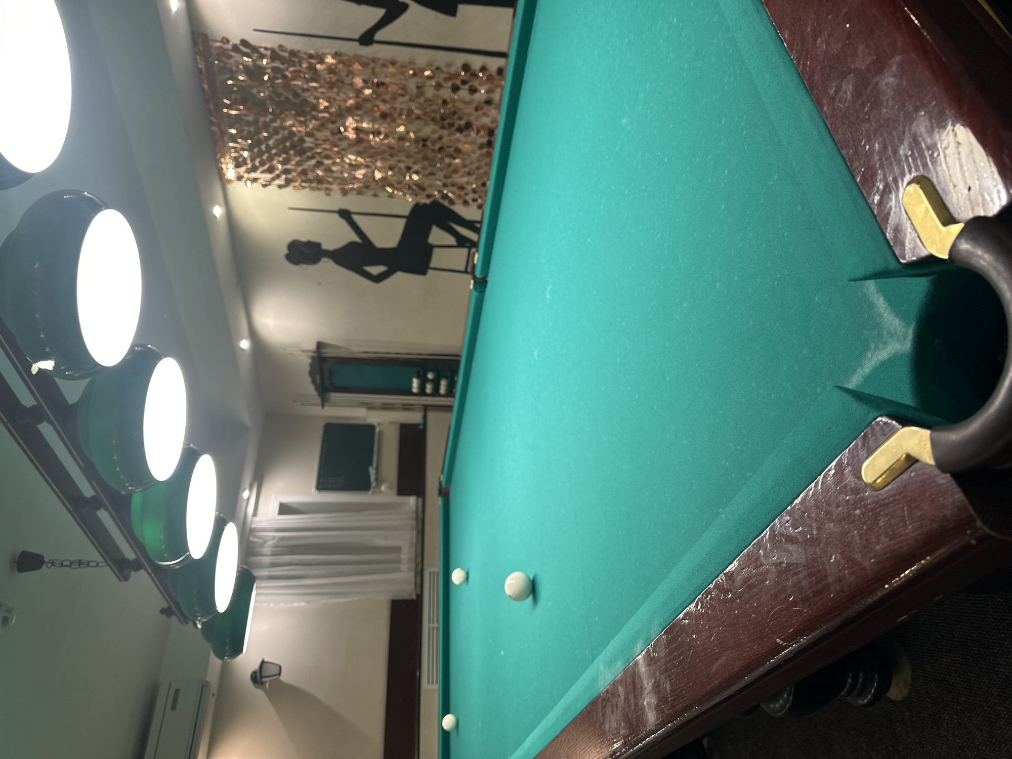 The image depicts a billiard table in a dimly lit room. The table is covered in green felt, with several white billiard balls scattered across its surface. The edge of the table is made of dark wood. Above the table, a row of green lamps hangs from the ceiling, illuminating the playing area. In the background, a wall is adorned with decorative golden streamers and silhouettes of people playing billiards. The room also features a window with sheer white curtains and what appears to be an air conditioning unit mounted on the wall. The overall ambiance suggests a recreational space. Based on the provided information, this scene is located in the city of Perm, Russia.