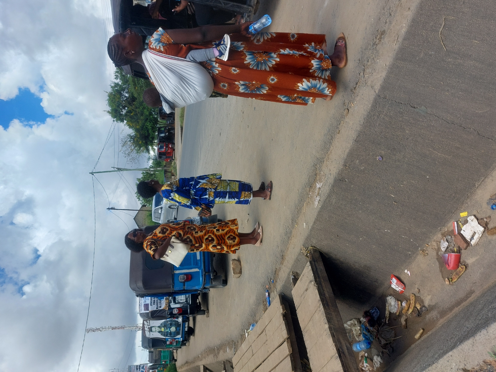 The image depicts a street scene in Dar es-Salaam, Tanzania. Three women are standing on the side of the road. One woman is carrying a baby in a white sling and holding a water bottle. She is wearing a brown floral dress. The other two women are standing nearby wearing traditional clothing. Parked on the side of the road behind the women are several blue auto rickshaws or "tuk-tuks". In the foreground is a concrete gutter filled with trash. The sky is overcast with patchy clouds.