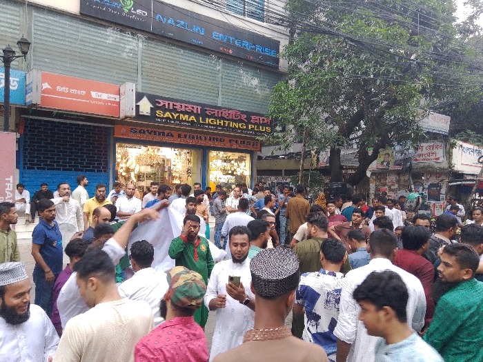 The image shows a densely packed crowd of people gathered on a street in Dhaka, Bangladesh, at the coordinates 23.734 latitude, 90.41 longitude. The street is lined with shops, including one with a sign that reads "Sayma Lighting Plus". The crowd is predominantly male, and many of them are wearing white shirts and traditional clothing. Some of the people are holding white flags. In the foreground, several men are huddled together, while others are standing around them, observing. One man in the center of the image is holding a mobile phone. There are also some people in the background, including a group of men standing in front of the "Sayma Lighting Plus" store. The street is narrow, and there are some trees and other buildings visible in the background. The street appears to be relatively calm and orderly, with no indication of any conflict or disturbance. The weather appears cloudy, with some rain possibly falling. The scene suggests a gathering of people in a crowded, urban environment.