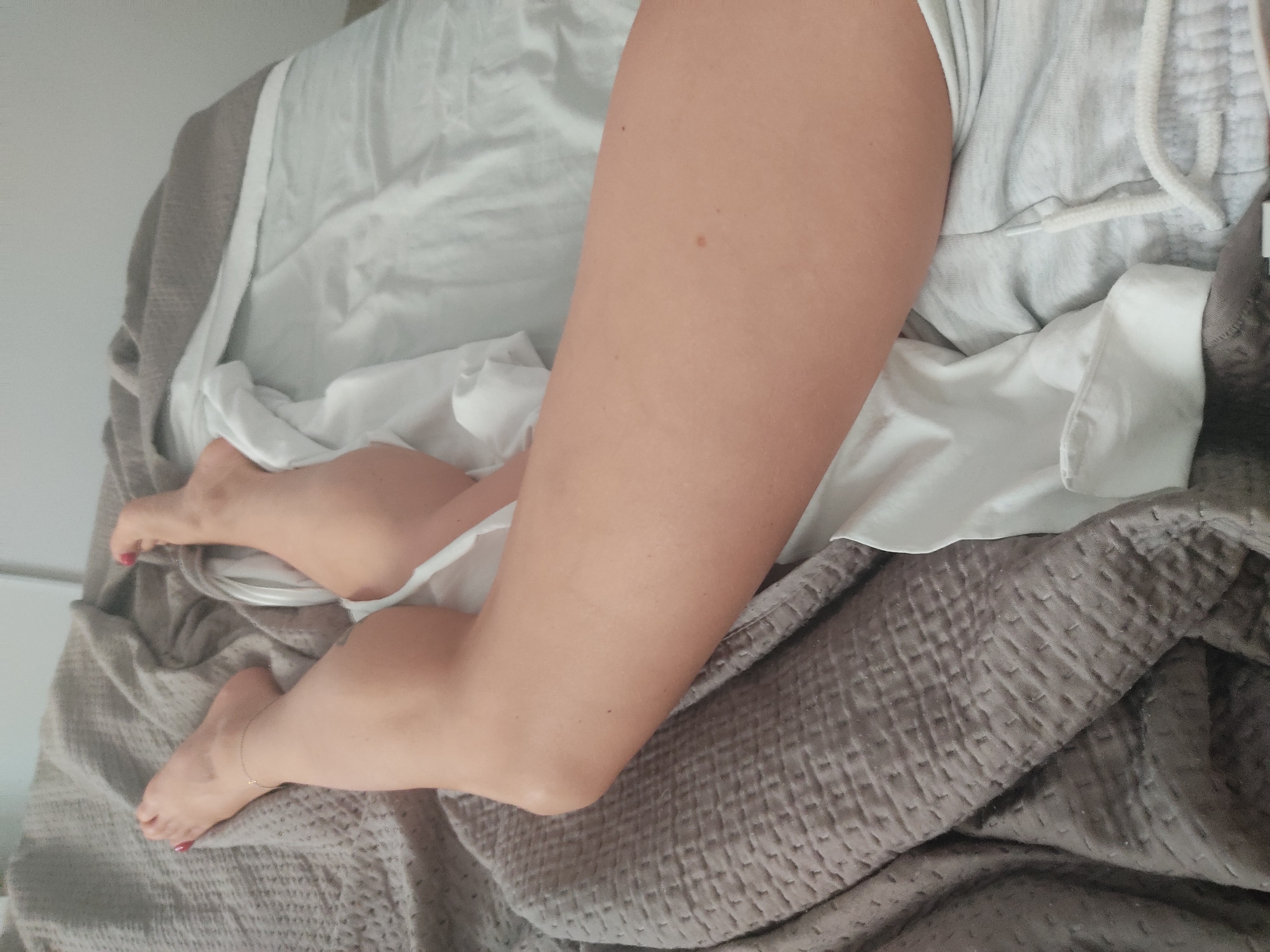 The image shows a close-up view of a person's legs and feet lying on a bed. The person has fair skin and is wearing red nail polish on their toenails. An anklet is visible on the left ankle. The bed is covered with a grey quilted blanket and white bedding. The scene appears to be indoors, possibly in a bedroom. No specific location in Blanes, Spain is identifiable from the image.