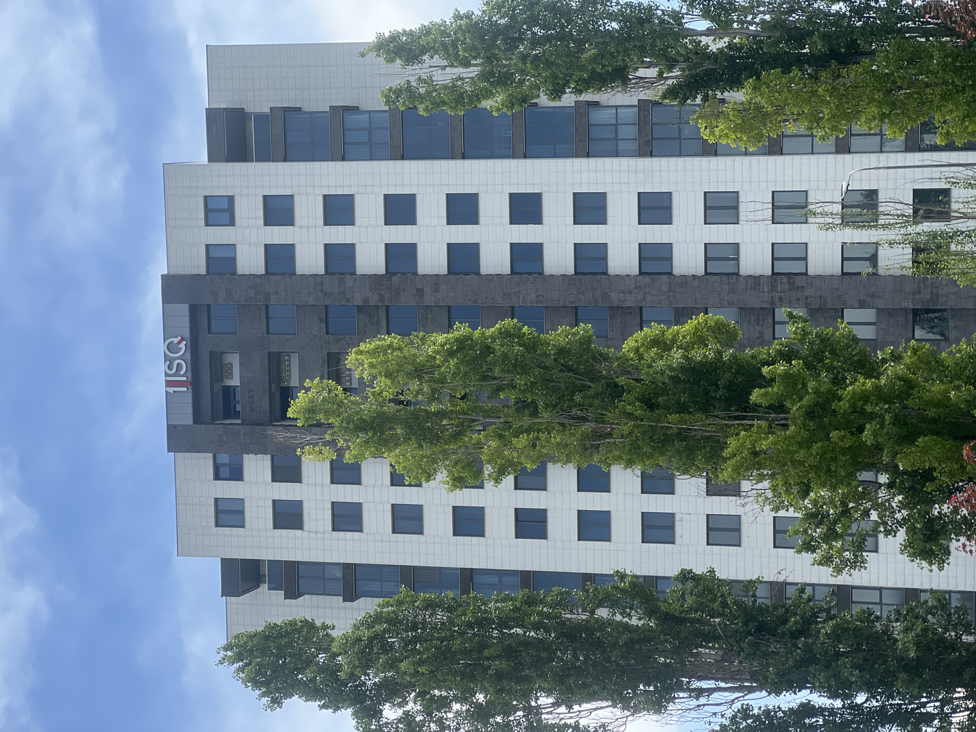 The image captures a tall building in Yerevan, Armenia, partly obscured by trees. The building's facade is a combination of white and gray tile, with rows of square windows. The building's name "ISQ" is vertically displayed on one side. The sky is blue with some clouds visible. The angle of the photo is slightly tilted, possibly from a ground-level perspective. Trees are situated in front of the building, partially blocking the view.