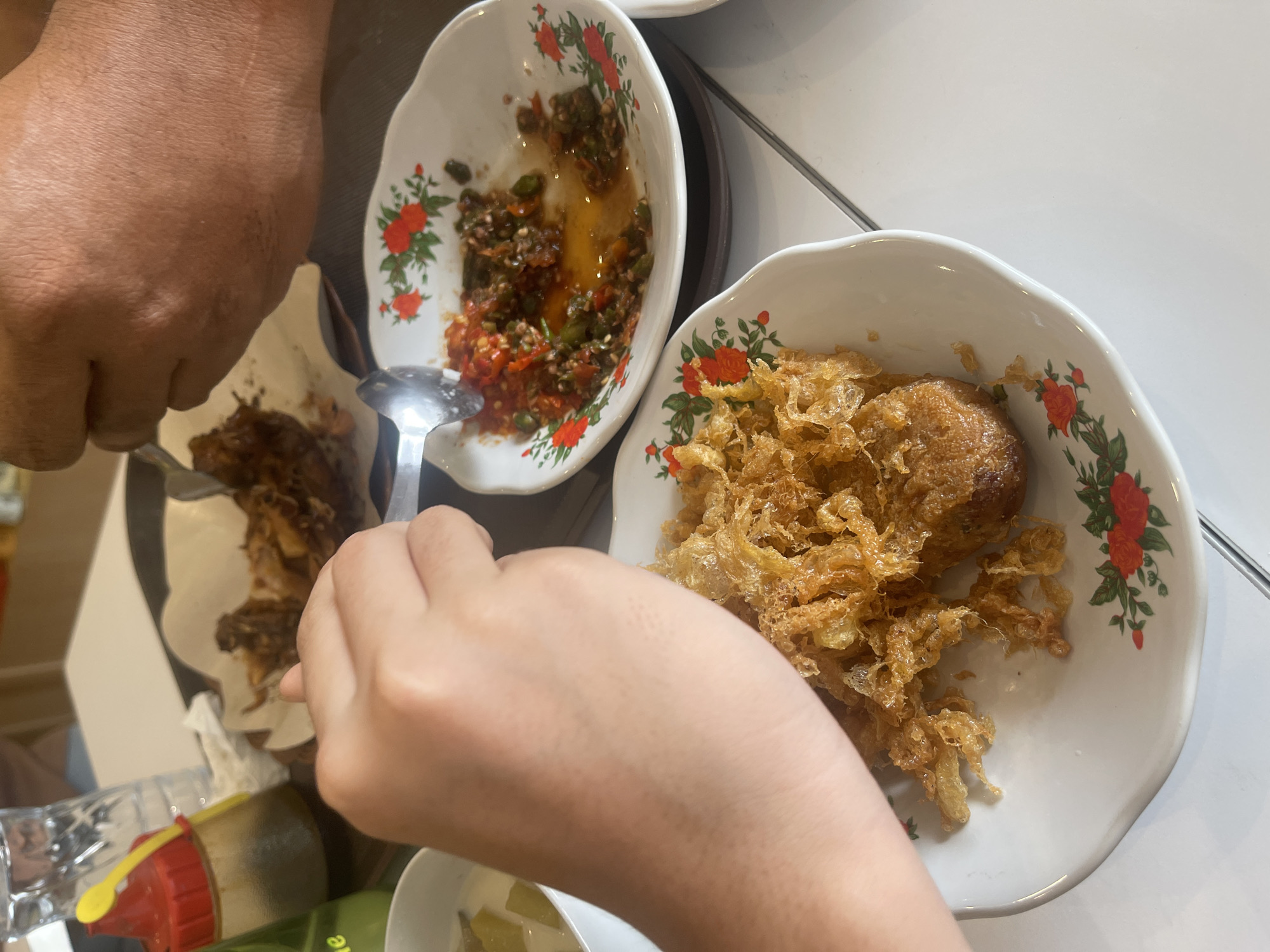 This is a close-up, overhead shot of a meal being shared at a table, likely in a restaurant in Medan, Indonesia. In the foreground, a person's left arm and hand are visible, holding a spoon. The spoon is hovering over a white plate filled with a golden-brown, crispy fried dish, possibly fried chicken or a type of fritter. The plate is decorated with small red and green floral patterns.
To the right of this plate is another white plate containing a vibrant red chili sauce or sambal, mixed with green chilies and oil. A metal spoon is partially submerged in this sauce. Further back, out of focus, is another dish of food, appearing to be fried meat, served on a piece of white paper. A clear glass of water and a condiment bottle with a red cap and yellow spout are also present on the table. The table surface is white, and the background is blurred, suggesting an indoor setting. The presence of sambal and the style of the fried food suggest it is Indonesian cuisine.
