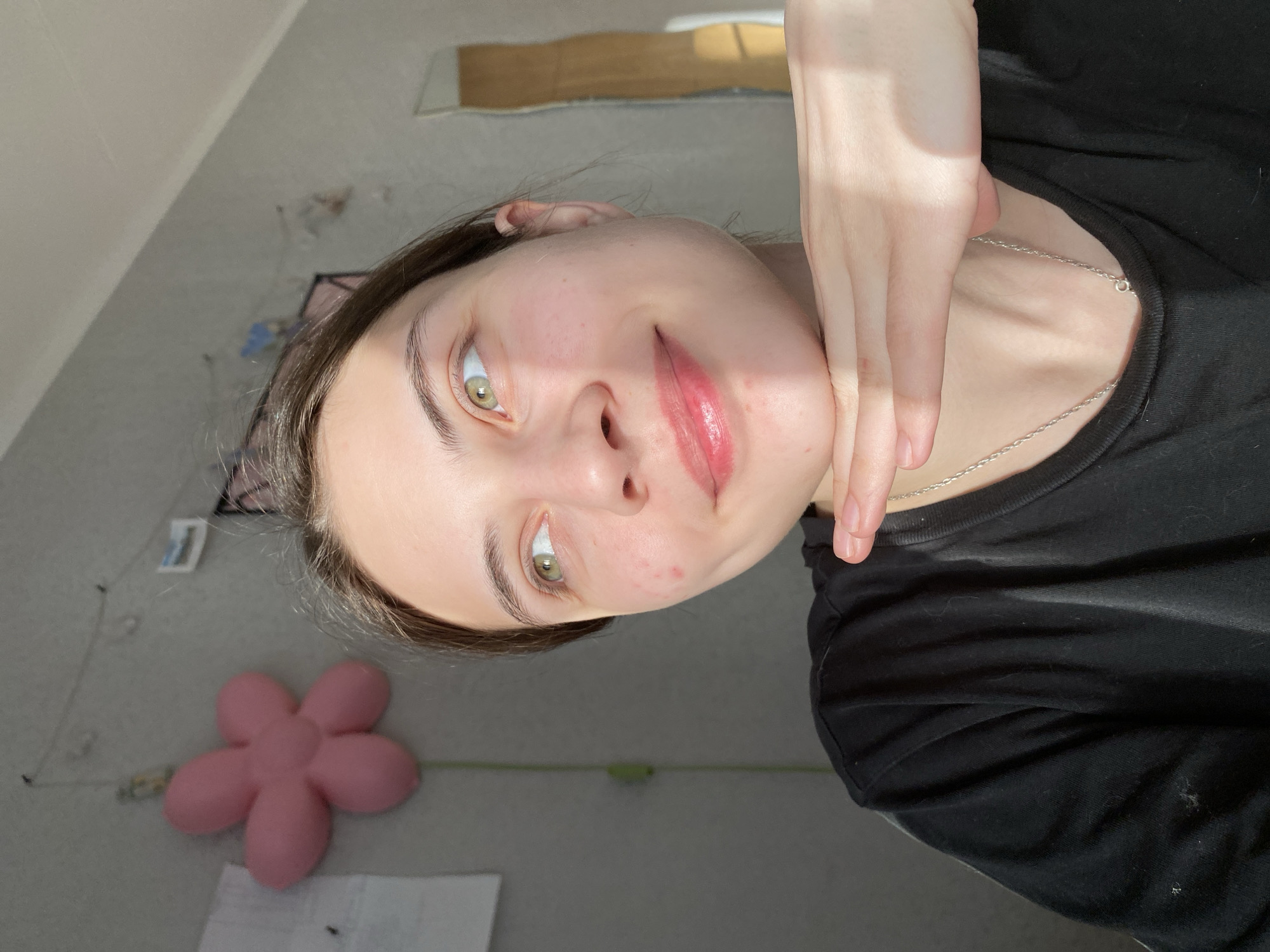 A woman with fair skin, green eyes, and dark brown hair is posing for a selfie. Her right hand is positioned under her chin. She is wearing a black t-shirt and a silver necklace. The background includes a grey wall with a pink flower-shaped pillow and a piece of paper. A window with sunlight streaming through can also be seen. The location cannot be determined from the visual information provided.