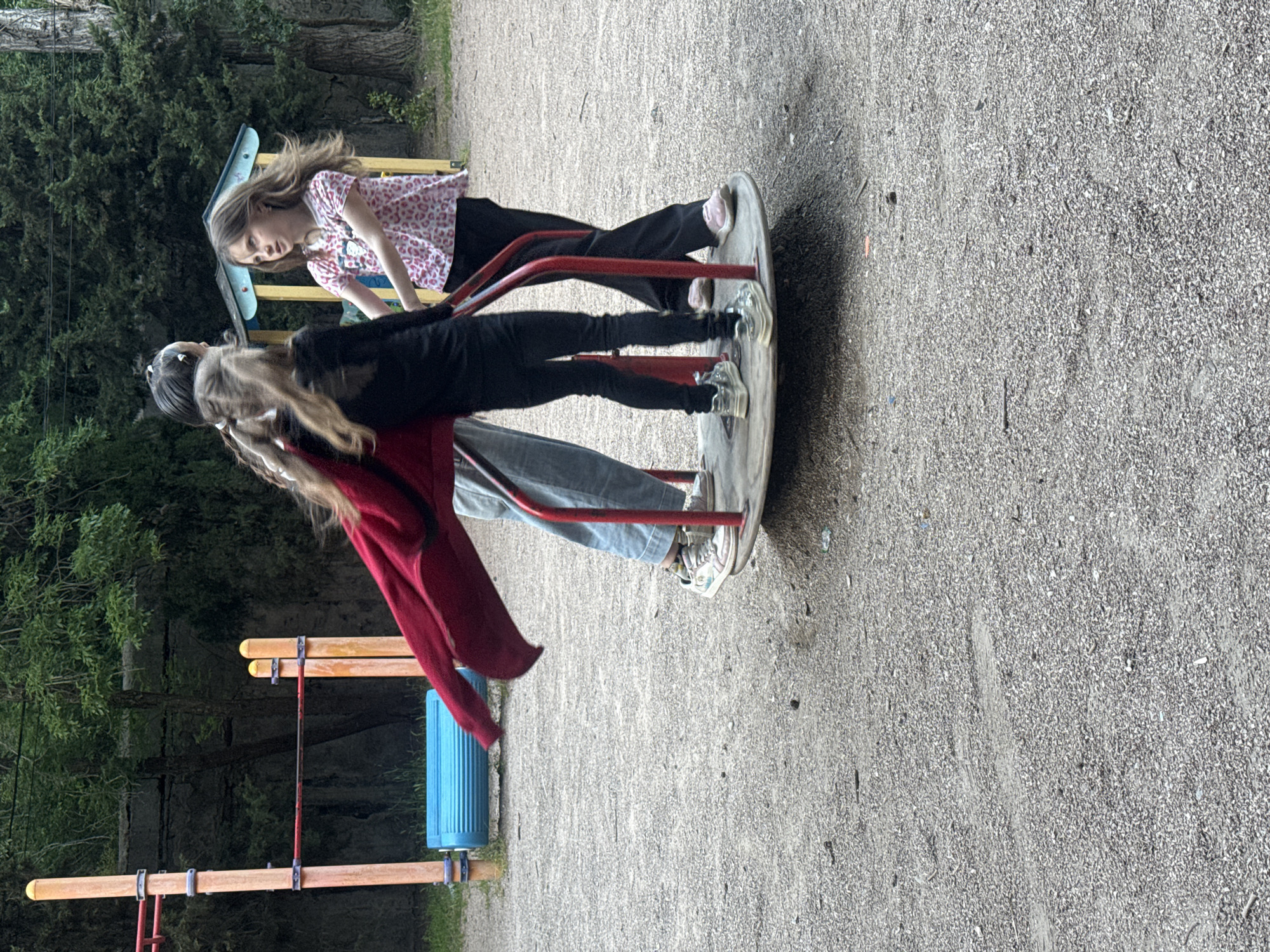 Three children are on a playground roundabout. One child, wearing a pink patterned shirt, black pants, and pink shoes, stands on the roundabout facing left. Another child, wearing a black shirt and pants with white shoes, stands facing right. A third child, wearing a red sweater and blue jeans, stands on the opposite side. The playground is located in a gravel area with green trees in the background and playground equipment to the side. The city and state are not identifiable.