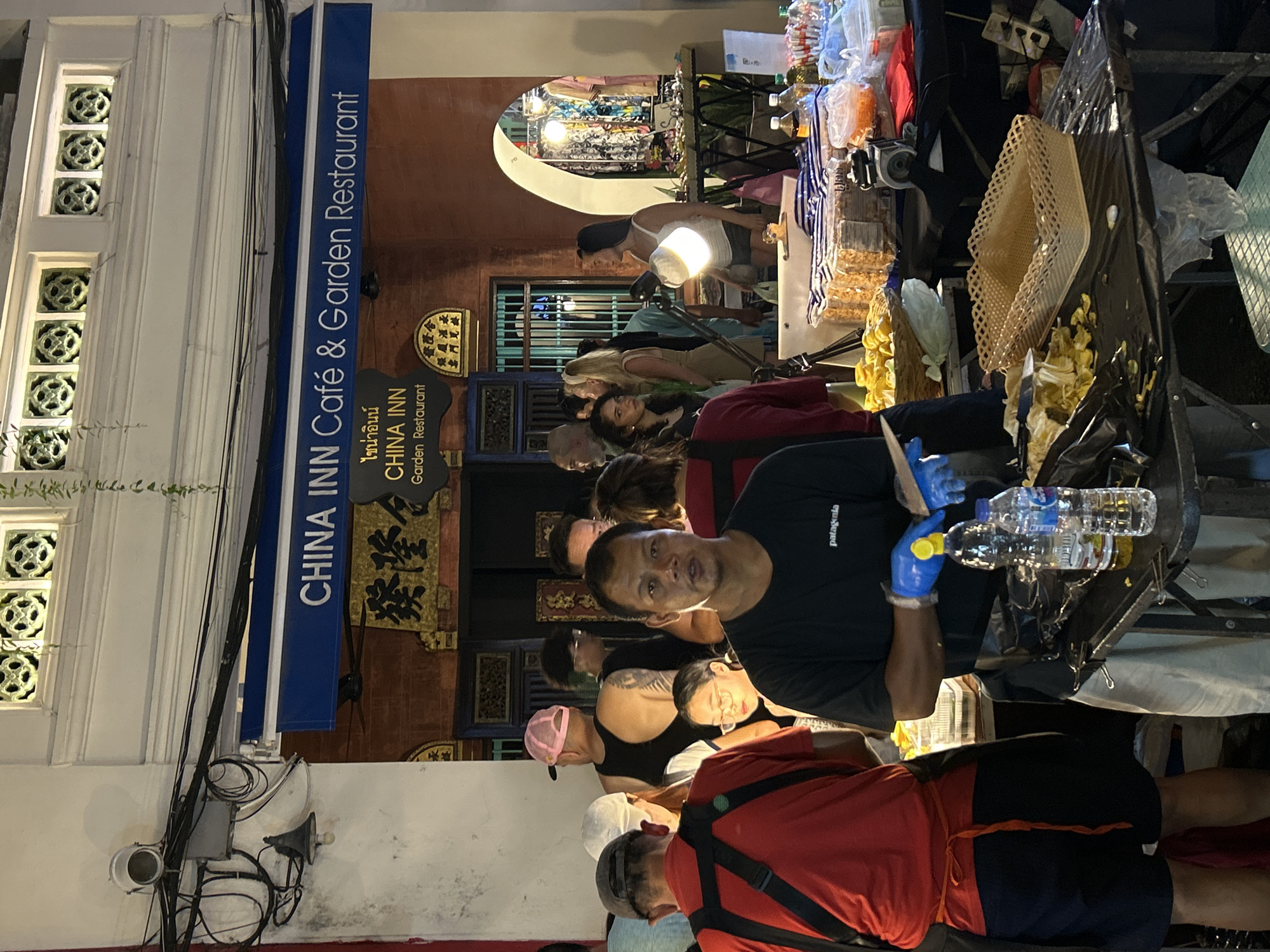In the city of Phuket City Municipality, Thailand, this image depicts a street scene at night, likely a market or a food stall area, in front of a restaurant identified as "CHINA INN Café & Garden Restaurant." A man in a black t-shirt and blue gloves is positioned in the foreground, facing the camera, appearing to be preparing food or handling items on a table covered with a black plastic sheet. Several bottles of liquid are on the table. To his left, another person in a red shirt and dark shorts is also visible, seemingly involved in food preparation or service. The background shows a crowd of people, some browsing or walking, and the facade of the restaurant with Chinese characters and the English signage. The overall atmosphere is bustling and suggests a commercial setting.