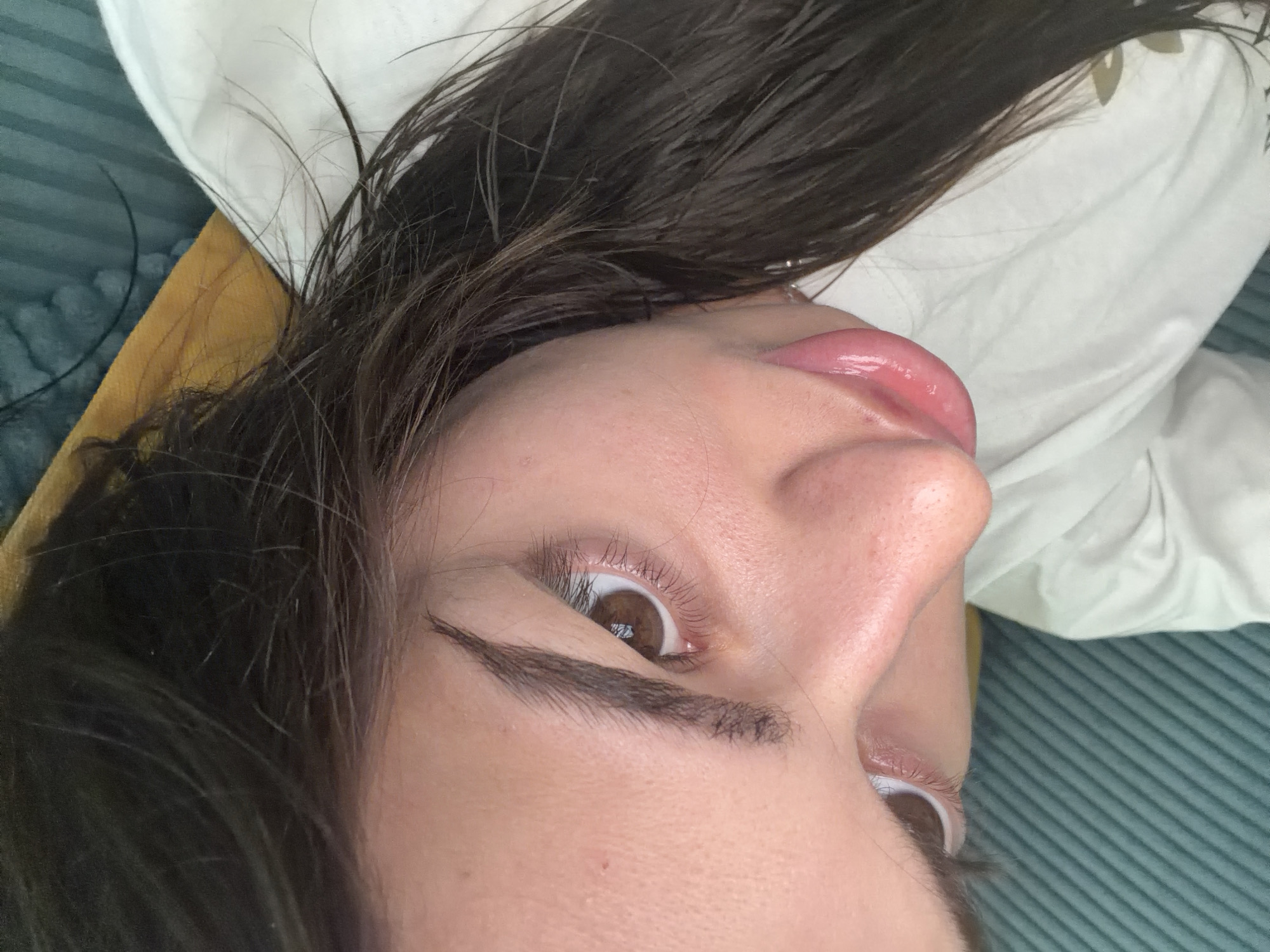 A close-up, high-angle shot focuses on the face of a person with brown eyes and dark brown hair. The person is wearing a white t-shirt and is lying down, possibly on a couch or bed, indicated by the textured blue fabric and a yellow pillow visible in the background. The lighting is soft, highlighting the person's facial features, including their nose and lips, which appear to have a glossy sheen. The background is blurred, drawing attention to the subject in the foreground.