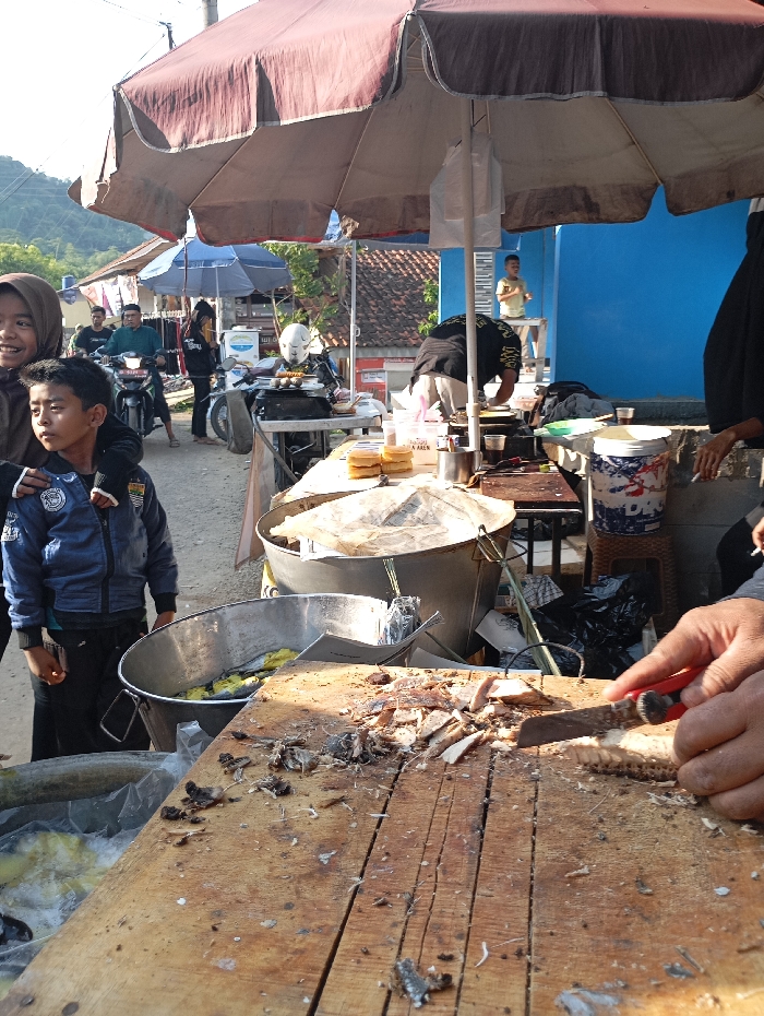 The scene is a food stall in Kecamatan Kutawaringin, Indonesia, located at coordinates -6.973 latitude and 107.52 longitude. A large, brown umbrella provides shade over the stall, which is situated in front of a blue-walled building. A wooden table, covered in a layer of fish bones and skin, is positioned under the umbrella. A man's hand, visible from the wrist down, is holding a red-handled knife and appears to be cutting up fish on the table. A large metal bucket with fish bones and skin inside sits beside the table. There is also a smaller bucket containing pieces of what appears to be fish and yellow-colored objects. Two young boys, one wearing a blue jacket and the other wearing a red shirt, stand near the table and watch the man cut the fish. Behind them, a man is riding a motorcycle, and two women are standing nearby. To the left of the table is a large, metal container, with an opening at the top. Another large metal container is visible to the left of the table. A third metal container sits on the table beside the man cutting the fish, with additional metal containers and other items behind it. A blue and white cooler sits on the table on the far right side of the image.