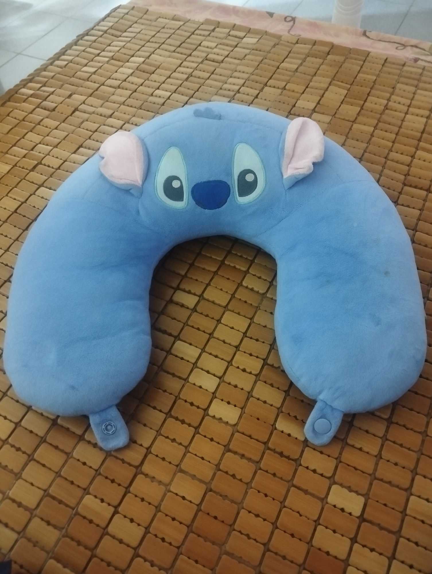 A blue Stitch character travel pillow is placed on a woven bamboo mat. The pillow has Stitch's characteristic large eyes, blue nose, and pink ears. It is shaped like a horseshoe, designed to support the neck. The bamboo mat is light brown and laid out in a grid pattern. In the background, a pink and white patterned surface is visible, and beyond that, a white cylindrical object. The scene appears to be indoors. There are no people or animals in the image. The lighting suggests it is daytime. There is no information in the image that indicates the location is Kaohsiung, Taiwan.