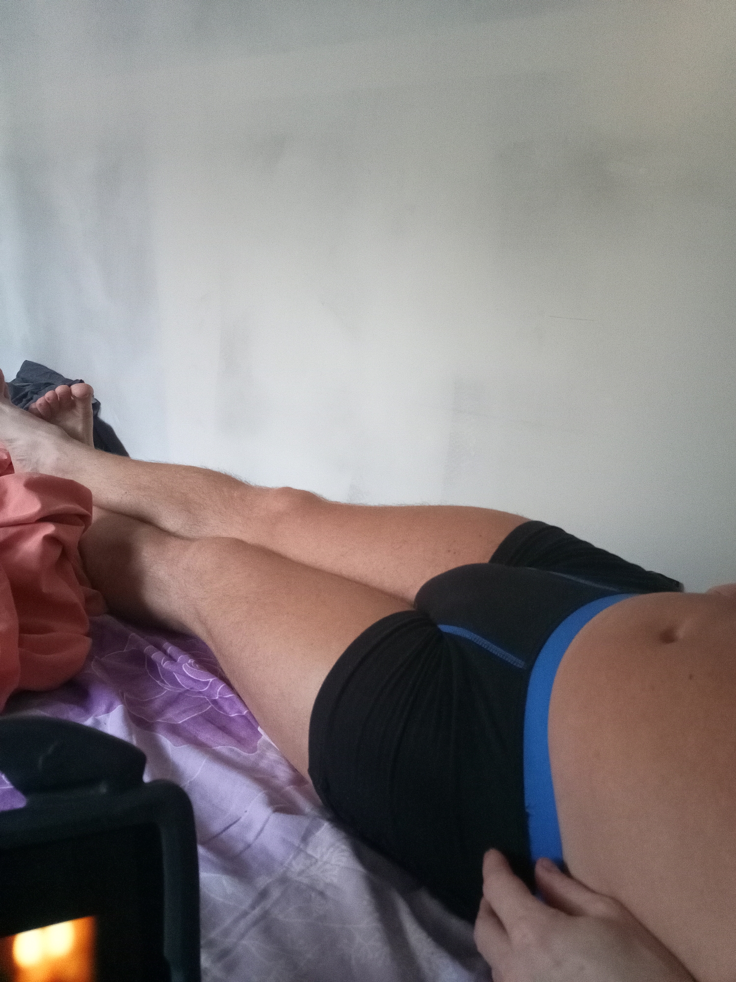 An individual is lying down on a bed. The person is wearing black boxer briefs with a blue waistband. Their legs are crossed. A pink garment and a dark gray garment are on the bed, near the person's legs. A tablet device is visible in the bottom left corner of the frame. The background is a plain white wall.