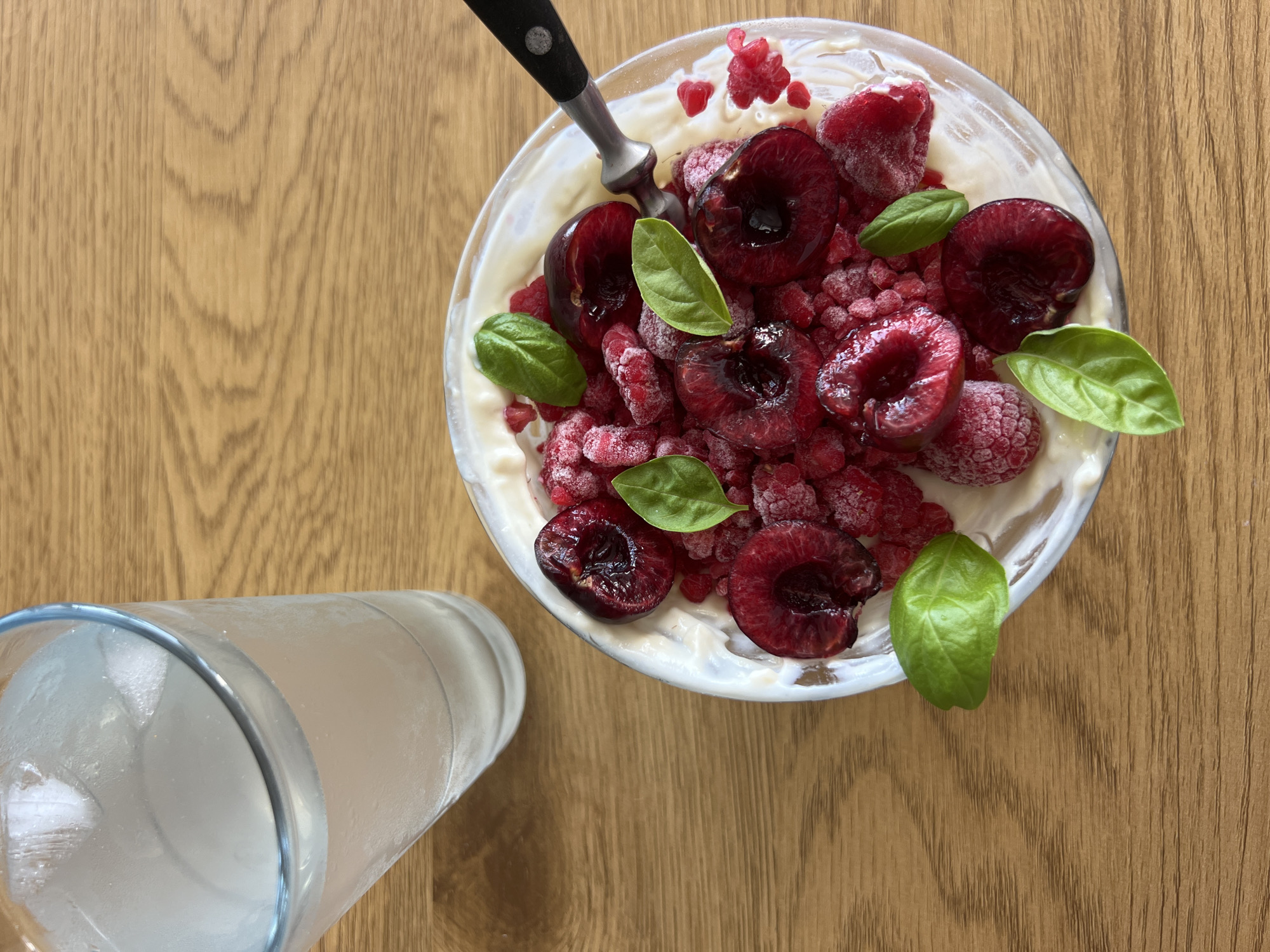 A close-up, overhead shot shows a bowl of yogurt topped with fresh cherries and raspberries, garnished with basil leaves. A silver spoon is partially submerged in the yogurt. To the left of the bowl, a tall glass of water, filled with ice and condensation, rests on a wooden table. The wooden surface exhibits a natural grain pattern and appears to be oak. The lighting suggests it is daytime, with no specific indication of weather conditions or time of day, though the natural light might imply it's not evening. The scene appears to be set indoors, likely in a kitchen or dining area, given the presentation of food and drink. No people are visible in the image. The overall impression is one of a fresh, healthy, and simple meal or snack.