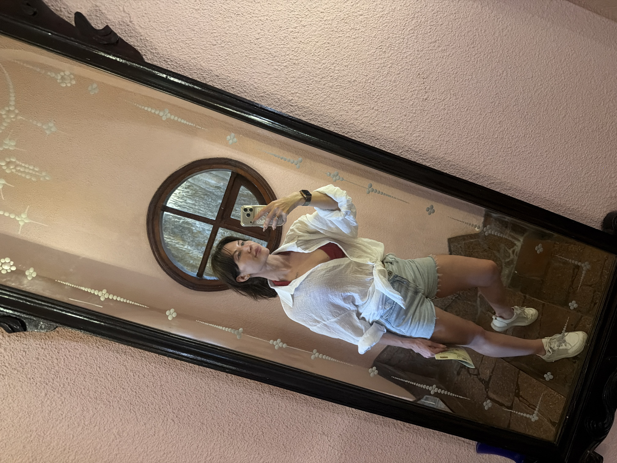 A woman stands in front of a large ornate mirror, taking a selfie. She is wearing a white unbuttoned shirt tied at the waist, a red bralette, denim shorts, and white sneakers. She is holding a yellow object in her left hand. The mirror reflects her and the surrounding environment, which includes a pink textured wall and a small circular window with wooden panes. The mirror itself has a dark wooden frame adorned with decorative elements. The floor in the reflection is tiled with brown and grey stones. The overall scene suggests an indoor setting, possibly a hotel room or a boutique, with a slightly vintage or bohemian aesthetic. The location is identified as Da Lat, Vietnam.