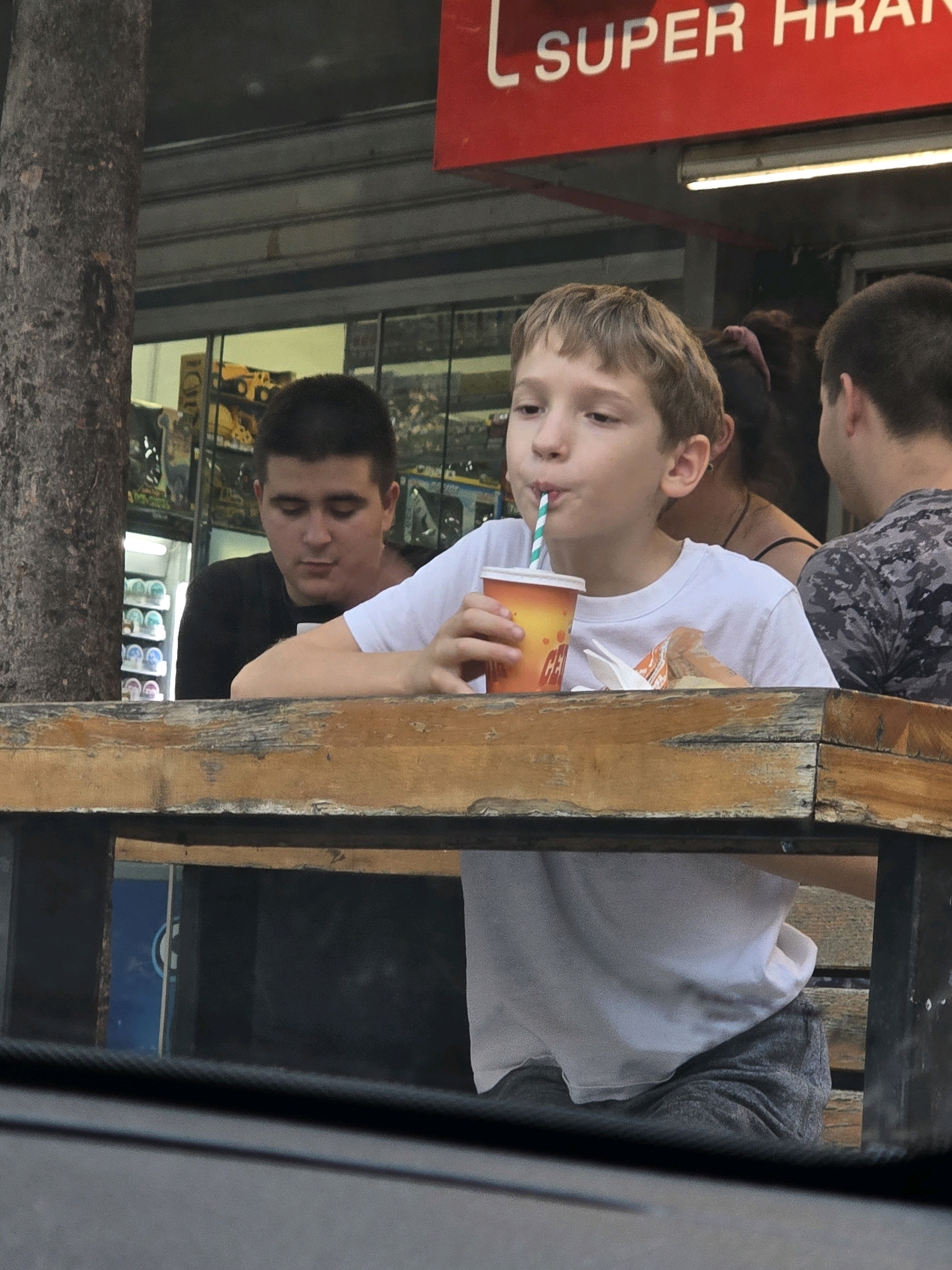 The image depicts an outdoor scene in front of a store with a red awning that reads "SUPER HRANA". A young boy wearing a white t-shirt and grey shorts is seated at a wooden table, drinking from a cup with a green and white striped straw. To his left, a young man with dark hair wearing a black t-shirt is also seated at the table, looking downwards. Behind them, a person with dark hair and a pink hair tie is partially visible. The table is made of rough, weathered wood, and behind the subjects, a store interior is visible with shelves containing various items. The scene appears to be in a public area, possibly a street or pedestrian walkway in Podgorica, Montenegro, given the context. The overall lighting suggests it is daytime.