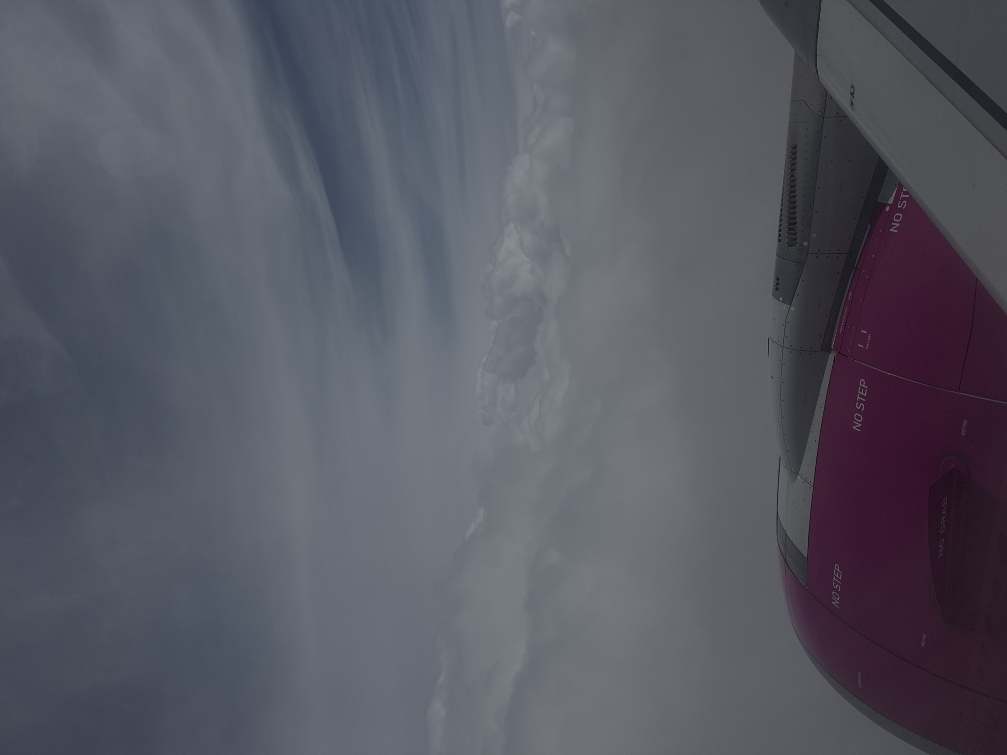 The image captures a view from an airplane window, looking down at a bright pink aircraft engine cowling with "NO STEP" markings visible. Above the engine, a layer of dense, white clouds obscures the view of the ground. Further up, wispy, grey clouds streak across a darker grey sky, suggesting an overcast or pre-storm condition. The image does not provide any information or visual cues to determine the location within the city of Warka, Poland.