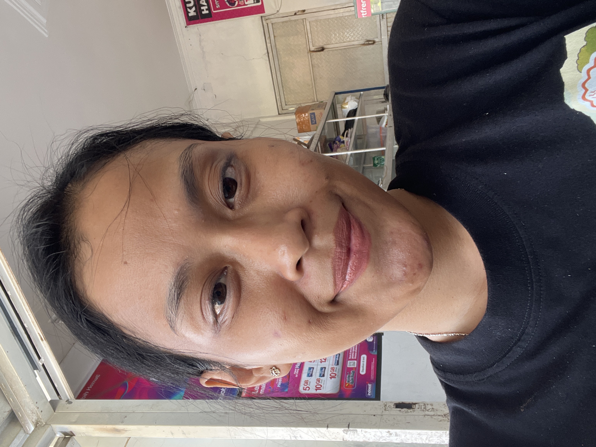 A close-up, head-and-shoulders selfie shows a young woman looking directly at the camera. She has dark, pulled-back hair and a light complexion with some blemishes on her face. She is wearing a black t-shirt and a delicate silver necklace. Her makeup is subtle, with lipstick in a muted pink shade. The background appears to be an outdoor or semi-outdoor location, possibly a shop or street stall, with signs and shelves visible. A poster with Indonesian text and graphics is partially visible on the left, and a doorway with frosted glass panels is seen in the background on the right. The overall lighting suggests it is daytime.