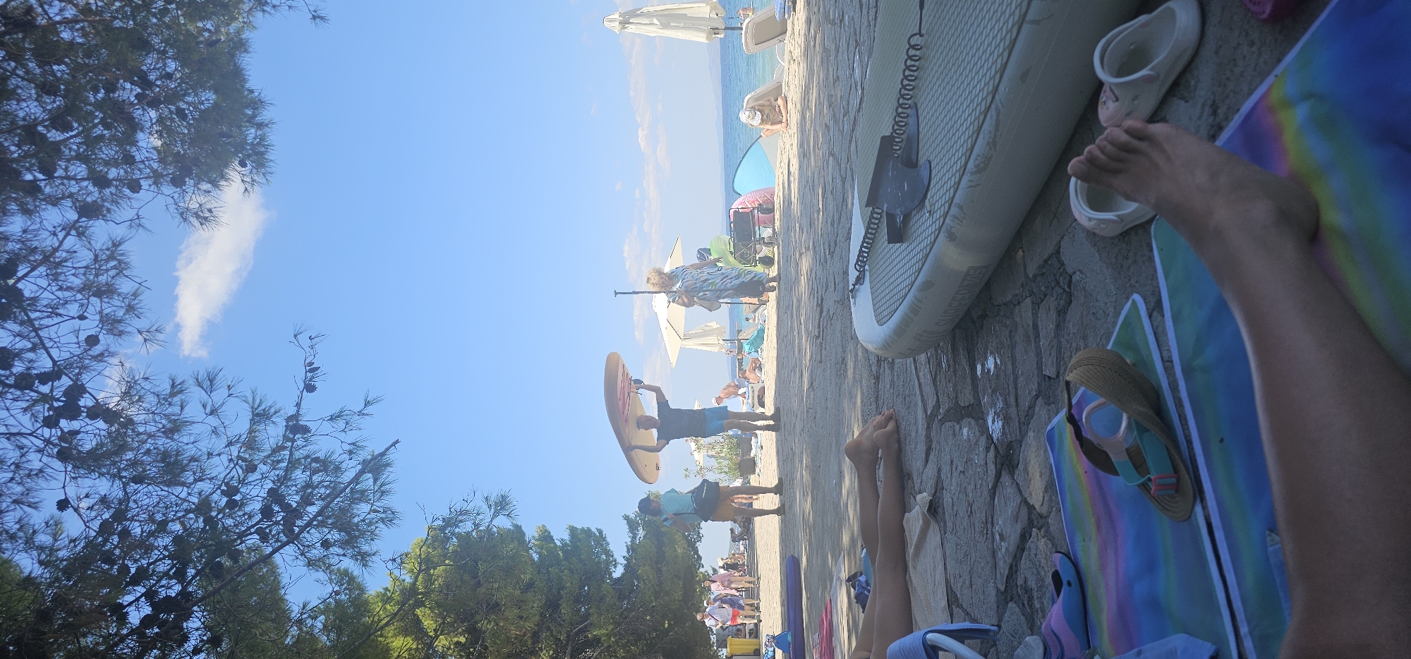 The image depicts a crowded beach scene in Grad Supetar, Croatia, under a clear blue sky with scattered clouds. The foreground is dominated by a person's legs and feet resting on colorful beach mats, with a paddleboard lying horizontally on a stone pathway. To the left, other people are visible lounging on mats and beach chairs, with a pair of flip-flops nearby.
In the middle ground, a man is carrying a paddleboard over his head, walking towards the right of the frame. A woman in a patterned dress is walking in the same direction, slightly behind him. To their left, a person in a light blue shirt and shorts stands with a bag, facing the water. Further back, numerous beach umbrellas and chairs are set up, with people sunbathing and relaxing. The beach itself is sandy, meeting the calm blue water of the sea. Trees with pine cones are visible on the left side of the image, providing shade over the pathway and beach. In the background, more people and beach amenities are scattered along the coastline, with a distant landmass visible across the water. The overall atmosphere suggests a busy summer day at the beach.