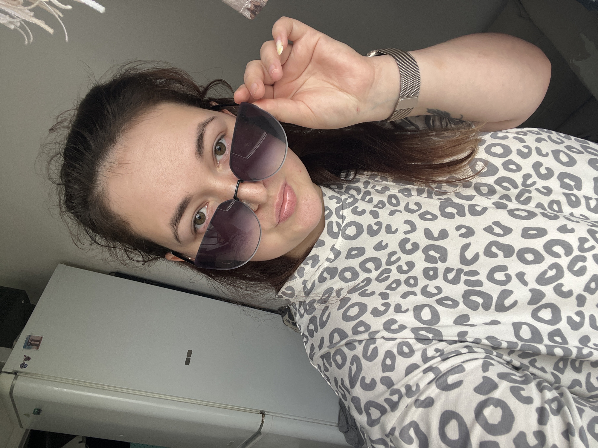 The image shows a young Caucasian woman with brown hair and green eyes, wearing large, dark-tinted sunglasses and a grey and white leopard-print shirt. She is holding the edge of her sunglasses with her right hand. A silver mesh watch is visible on her left wrist. To her left is a white refrigerator, possibly in a kitchen or utility area. The background is a plain, grey wall. There is no indication of the city of Chelyabinsk, Russia, in the image.