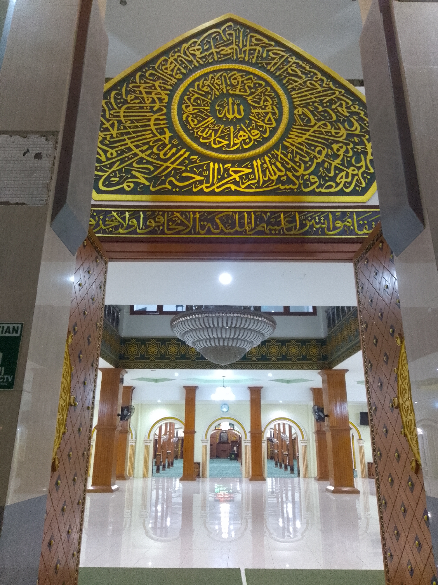 The image depicts the interior of the Alun-Alun Mosque in Lamongan, Indonesia. The foreground is dominated by an ornate wooden doorway with inlaid geometric patterns and gold accents. Beyond the doorway, the spacious prayer hall is visible, characterized by rows of polished wooden columns supporting arched doorways. The ceiling features a large, elaborate chandelier. Above the entrance to the prayer hall, a large, decorative panel displays intricate Arabic calligraphy in gold lettering on a dark green background. The floor is a highly reflective white tile, creating mirror-like reflections of the architectural elements. The overall impression is one of grandeur and religious significance. There are no people visible in the image.