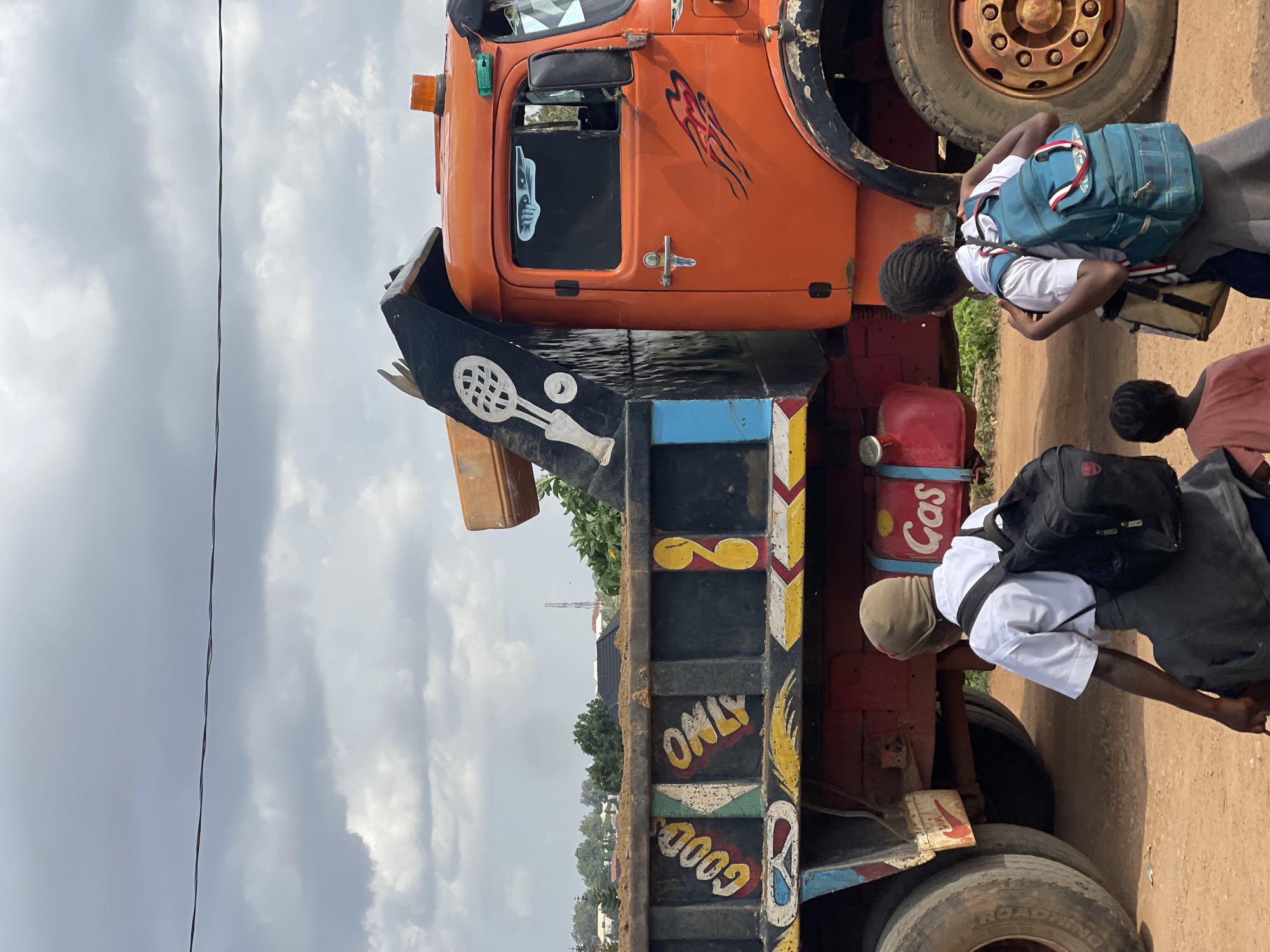 A vibrant, slightly hazy image captures a scene on a dirt road in Jos, Nigeria, under a dramatic, cloudy sky. Dominating the frame is a large, orange dump truck with significant wear and tear, its side adorned with colorful hand-painted designs and text. The words "GOOD ONLY" are prominently displayed in yellow letters within painted panels. A black depiction of a tennis racket and ball is visible on the truck's raised bed. To the right of the truck, a bright red gas tank is mounted, also featuring painted text.
In the foreground, three individuals are walking away from the viewer. The two closest are young girls wearing school uniforms consisting of white shirts and grey skirts. Both have backpacks, one black and the other blue, slung over their shoulders. A smaller child, also wearing a pink shirt, is positioned slightly behind and between them. The ground is a dusty, reddish-brown dirt. In the background, a line of trees and buildings suggests an urban or semi-urban environment. A cell phone tower is visible in the distance. The overall impression is one of everyday life unfolding against a backdrop of both vibrant local artistry and the practicalities of transportation in the region.