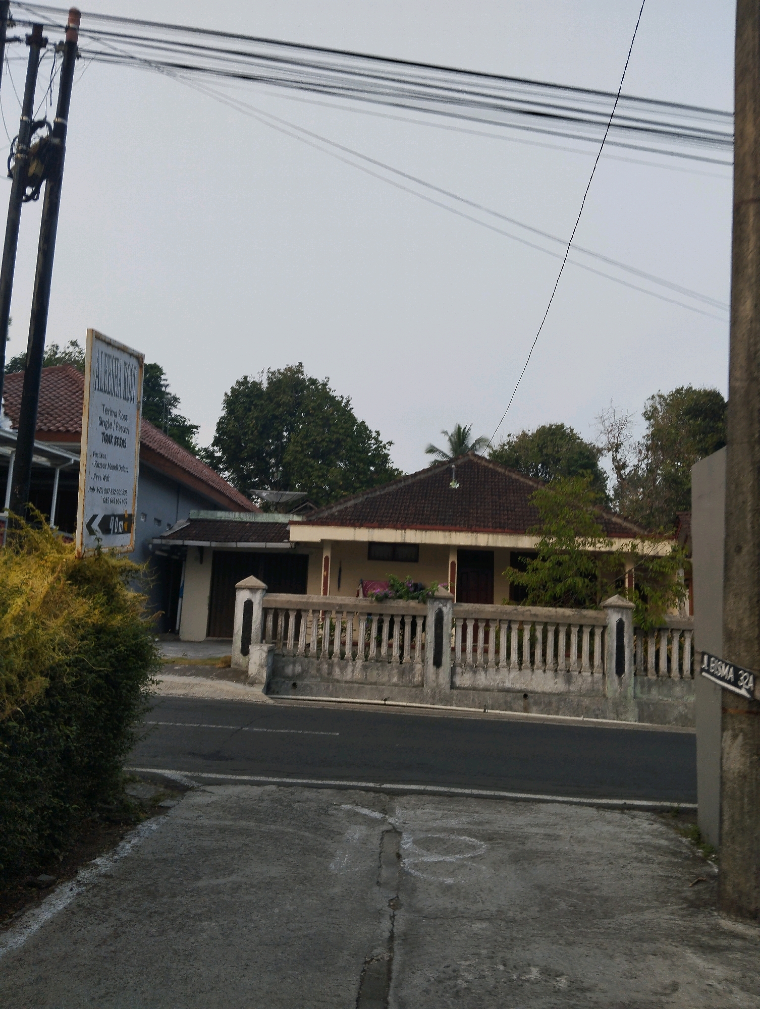 A medium shot captures a street scene in Salatiga, Indonesia, under an overcast sky. In the foreground, a concrete path leads toward the street, bordered by a sparse, yellow-green bush on the left. The street itself is paved and dark, with a white line marking its edge. To the right of the street, a low, ornate concrete fence with vertical bars runs along the property line of a house. The house, visible behind the fence, is a single-story structure with a reddish-brown tiled roof. A signpost with an attached directional sign reading "ALFESHA KOST" and further details about lodging is positioned to the left of the house. The background consists of trees and the sky. A utility pole with wires is prominent on the left side of the image, and another similar pole is visible on the far right. A street sign indicating "BISMA 32A" is attached to the right-hand pole.