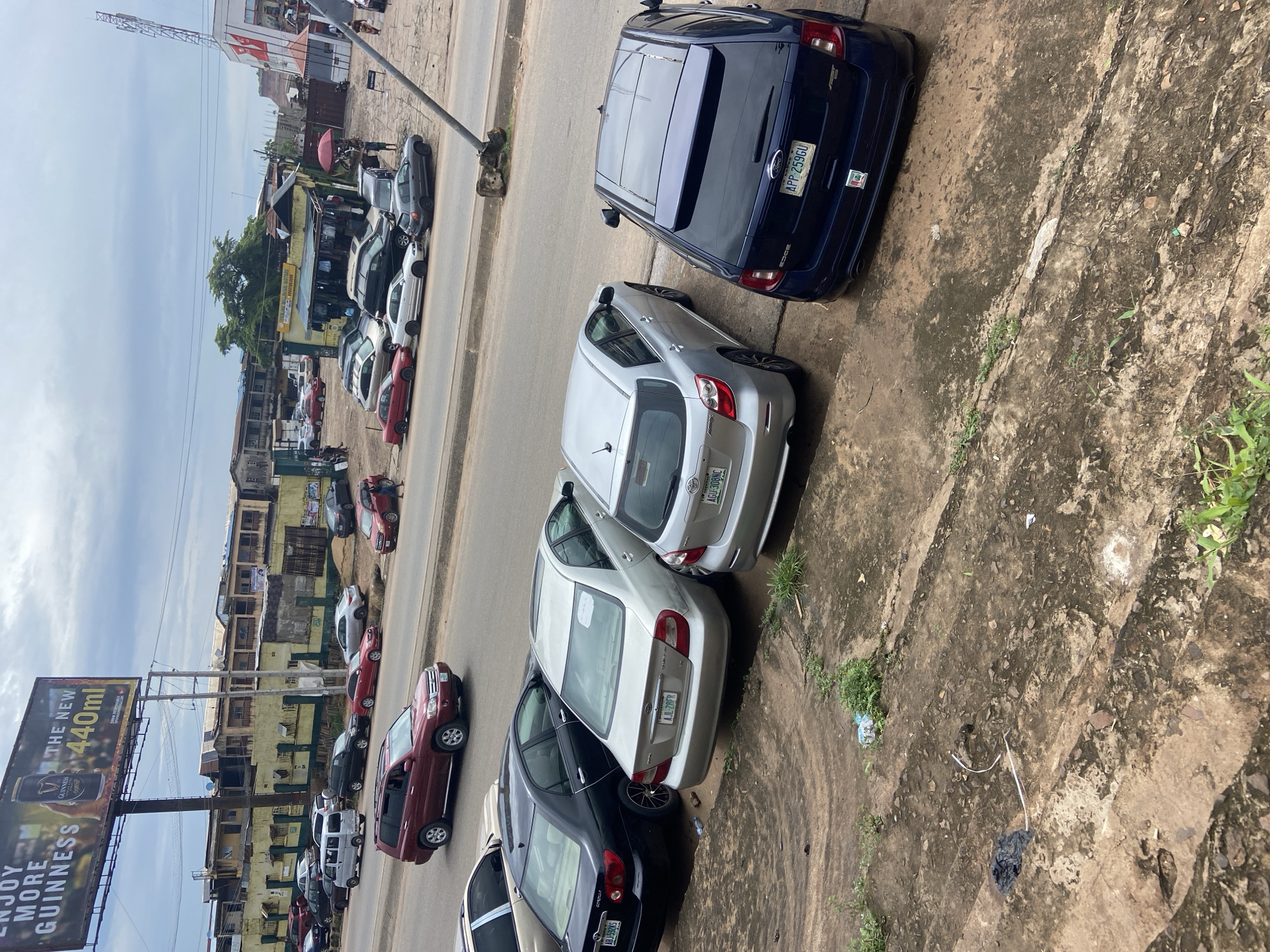 This image captures a street scene in Ogbunike, Nigeria, under an overcast sky. The foreground shows a gritty, concrete surface with sparse vegetation and scattered debris, including plastic bags and discarded items. A dark blue Ford Edge SUV is parked in the foreground, its rear facing the viewer, displaying a license plate that reads "APP 259GU." To its left, a silver Toyota Matrix is parked, also with its rear facing the camera, showing a license plate "AGU 308NC." Further left, a silver Toyota Camry with license plate "AJL 28FR" is parked next to a black Toyota Corolla with license plate "ABJ 592KS." Beyond these parked vehicles, a multi-lane road is visible with several other cars, including a red SUV and a white van, in motion or parked. The background features a row of buildings with weathered facades, some appearing to be shops or residential structures, along with a prominent billboard advertising Guinness beer. The overall impression is of an active urban environment with evident signs of wear and tear.