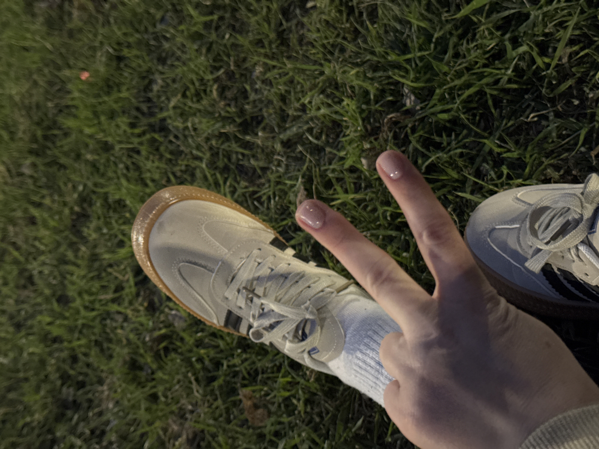A high-angle, close-up shot captures a person's left foot and hand on green grass, likely in Chaikovsky, Russia. The individual wears a light-colored, possibly off-white or light gray, sneaker with dark stripes on the side and a distinctive gum-colored sole. White ribbed socks are visible above the shoe. The person's left hand is raised in a "peace" sign, with the index and middle fingers extended. The fingernails are manicured with a glossy, light pink or nude polish.
The scene is dimly lit, suggesting it is evening or night, with artificial light sources creating highlights on the shoe and hand, and casting soft shadows. The grass appears dense and green, though its specific texture is softened by the low light. No distinct objects or structures are visible in the background, focusing attention solely on the foot, hand, and immediate ground. There is no visible text in the image. The overall mood is casual and slightly playful, conveyed by the hand gesture.