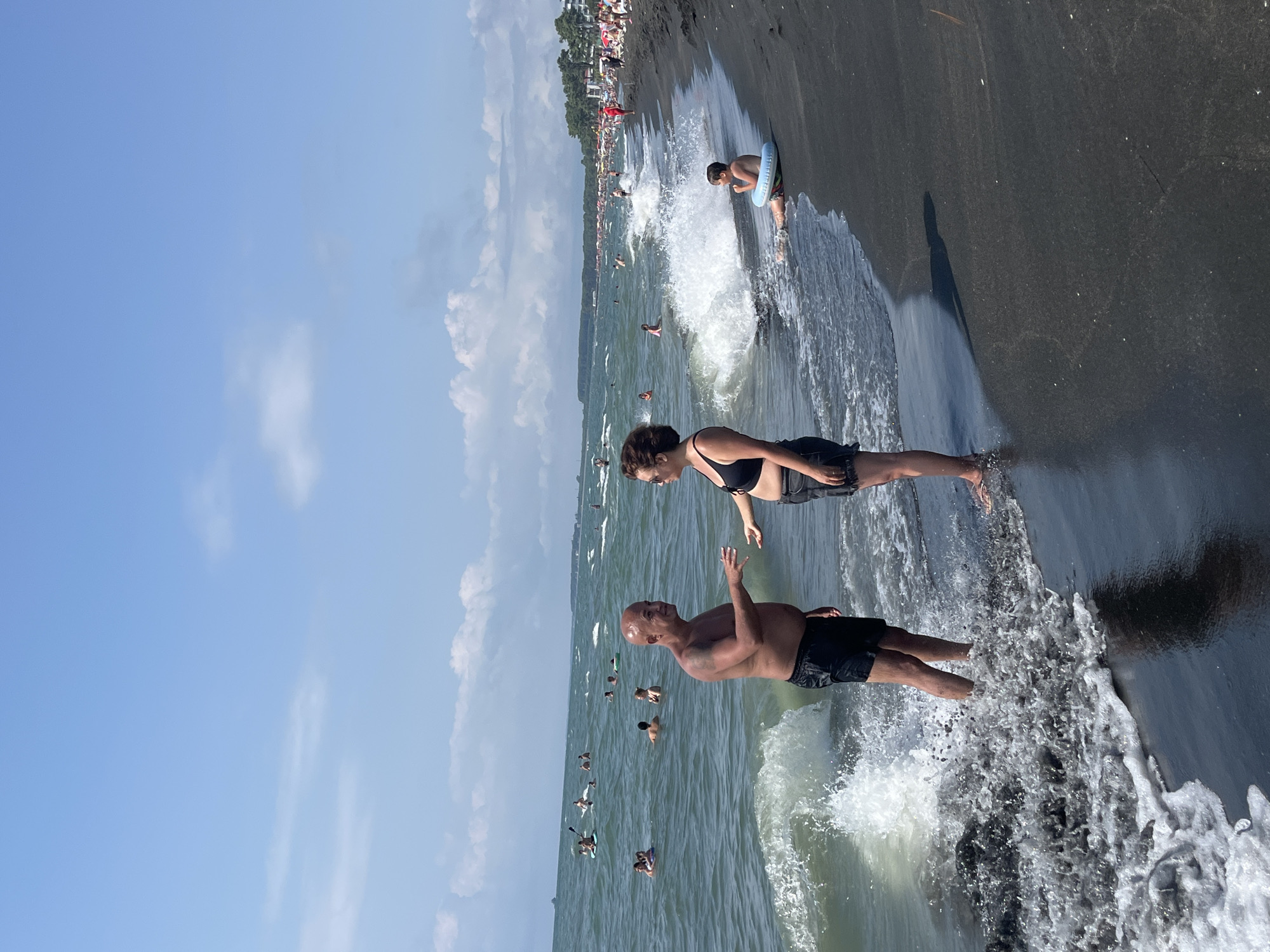 In Ureki, Georgia, a beach scene unfolds under a partly cloudy sky. In the foreground, a man wearing black swim trunks stands in the shallow water, facing a woman in a black bikini. The man has his arms extended, seemingly in mid-conversation. The woman, facing him, also has her arms out. Waves crash around their legs, with dark, volcanic sand visible on the beach.
Further into the water, numerous people are enjoying the sea. Some are swimming, while others are on inflatable devices or paddleboards. A child is seated on the shore to the right of the main couple, wearing a blue inflatable ring. In the background, a line of trees separates the beach from a developed area with buildings and umbrellas, suggesting a resort town. The sea is a moderate blue-green color, with whitecaps indicating some choppiness. The overall atmosphere is one of leisure and summer activity on a Georgia coastline.