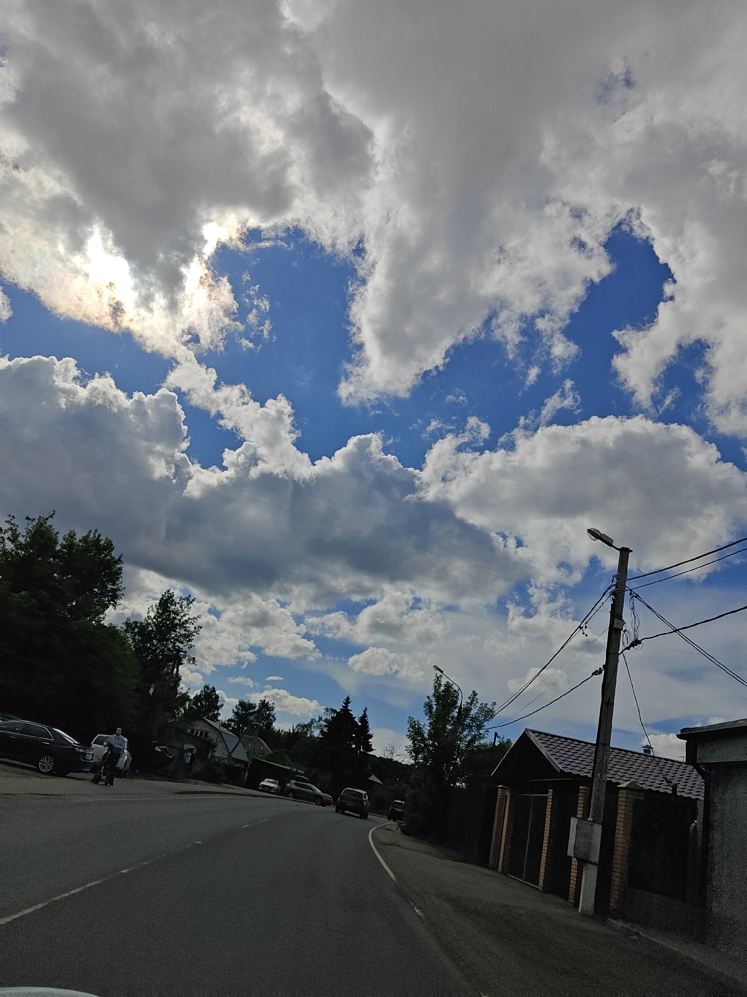 A low-angle, wide shot shows a paved road curving to the right, with a cloudy, bright blue sky overhead. The sun peeks through the clouds, creating a halo effect. On the left side of the road, several cars are parked, and a person on a scooter is riding past them. Behind the cars and scooter, there are trees and houses. On the right side of the road, there is a street light, power lines, and a house with a dark-tiled roof and a brick fence. The scene appears to be in a residential area, possibly a village or suburban neighborhood. The lighting suggests it is daytime, with a mix of sun and clouds indicating fair weather. There is no visible text in the image.