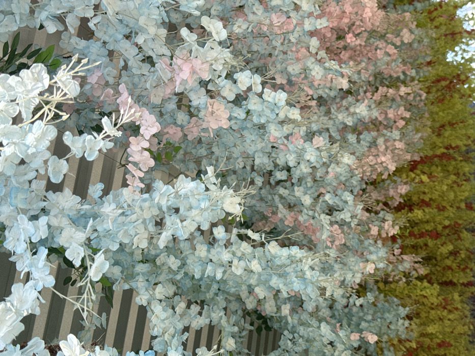 A cascade of pastel blue and pink blossoms drapes a wall. The wall behind features vertical grey and white stripes. To the right, a dense backdrop of verdant foliage with yellow undertones suggests a lush garden or forest. The focus is on the delicate floral arrangement, creating a serene and whimsical atmosphere.