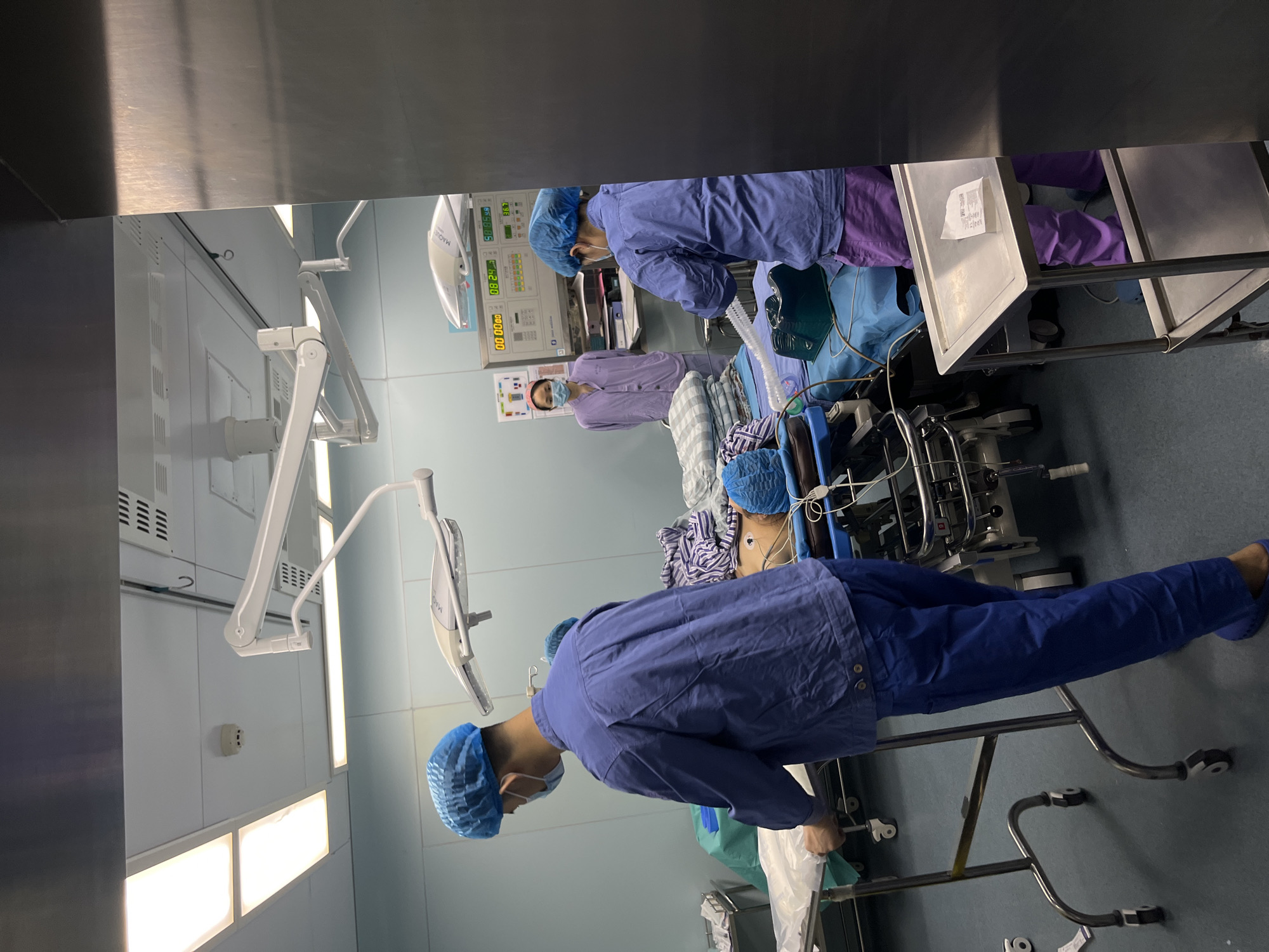 A surgical operating room in what is likely a hospital in Yuzhong District, China. Several individuals in blue surgical scrubs and caps are attending to a patient lying on an operating table. The room has medical equipment, lighting, and monitors visible. One individual is standing near the patient, adjusting equipment or attending to the patient's needs.