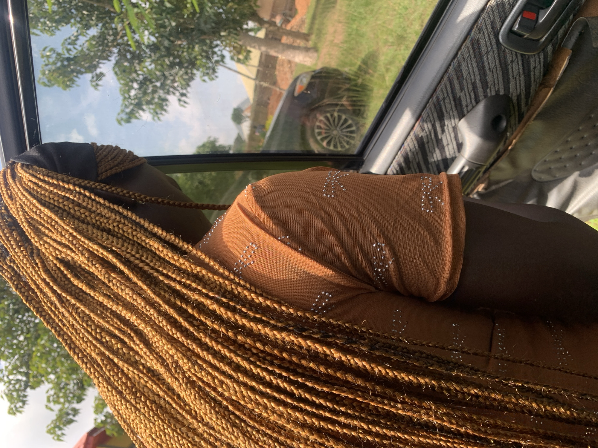 The image shows a young Black female from the shoulders up, viewed from behind and to the side, as she looks out the window of a car. She has long, golden-brown braided hair that cascades down her back and shoulders. She is wearing a sheer, burnt orange top adorned with small, scattered silver rhinestones. Her hair is partially covered by a black headscarf. The car's interior is visible in the foreground, with a patterned door panel and a black armrest. Through the car window, a blurry outdoor scene is visible, featuring green grass, trees, a dark-colored car, and buildings in the background, suggesting a semi-urban or rural setting. The lighting indicates it is daytime, with a bright sky visible through the trees.