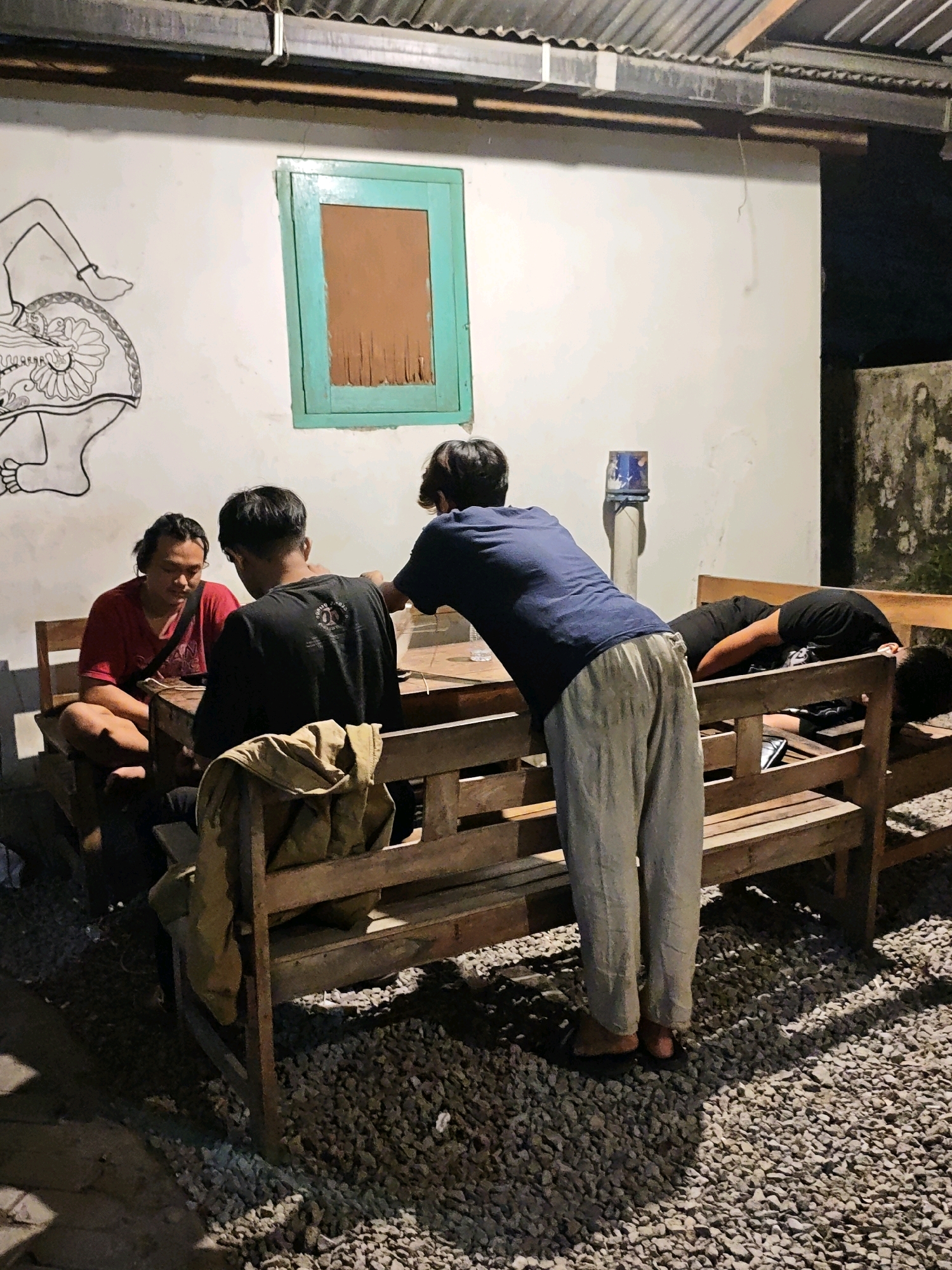 The image depicts an outdoor scene at night, likely in Ngasem, Indonesia. Four individuals are present, seated and standing around a wooden table and benches. On the left, a person in a red shirt is seated. To their right, another person in a black shirt is also seated. A third individual, wearing a blue t-shirt and light-colored pants, stands leaning over the table, facing away from the camera. On the far right, a fourth person is lying down on the bench, their head on their arm, with their body obscured by the bench. A tan-colored jacket or cloth is draped over the back of the bench in the foreground. The environment includes a white wall with a turquoise-framed window and a black-and-white mural on the left. The ground is covered in gravel, and a corrugated metal roof is visible overhead. The lighting suggests artificial illumination, casting shadows.