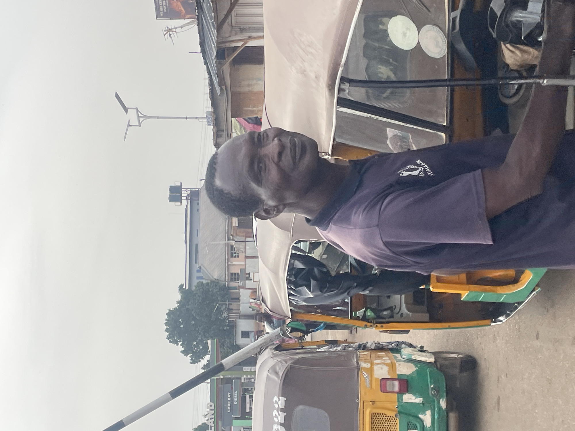 The image depicts an individual in Jos, Nigeria, standing next to a yellow and green auto-rickshaw. The person is wearing a purple shirt with a logo on the upper left side. The auto-rickshaw is in a street environment with buildings, a streetlight, and other commercial signs in the background, including what appears to be a gas station. The sky is overcast.